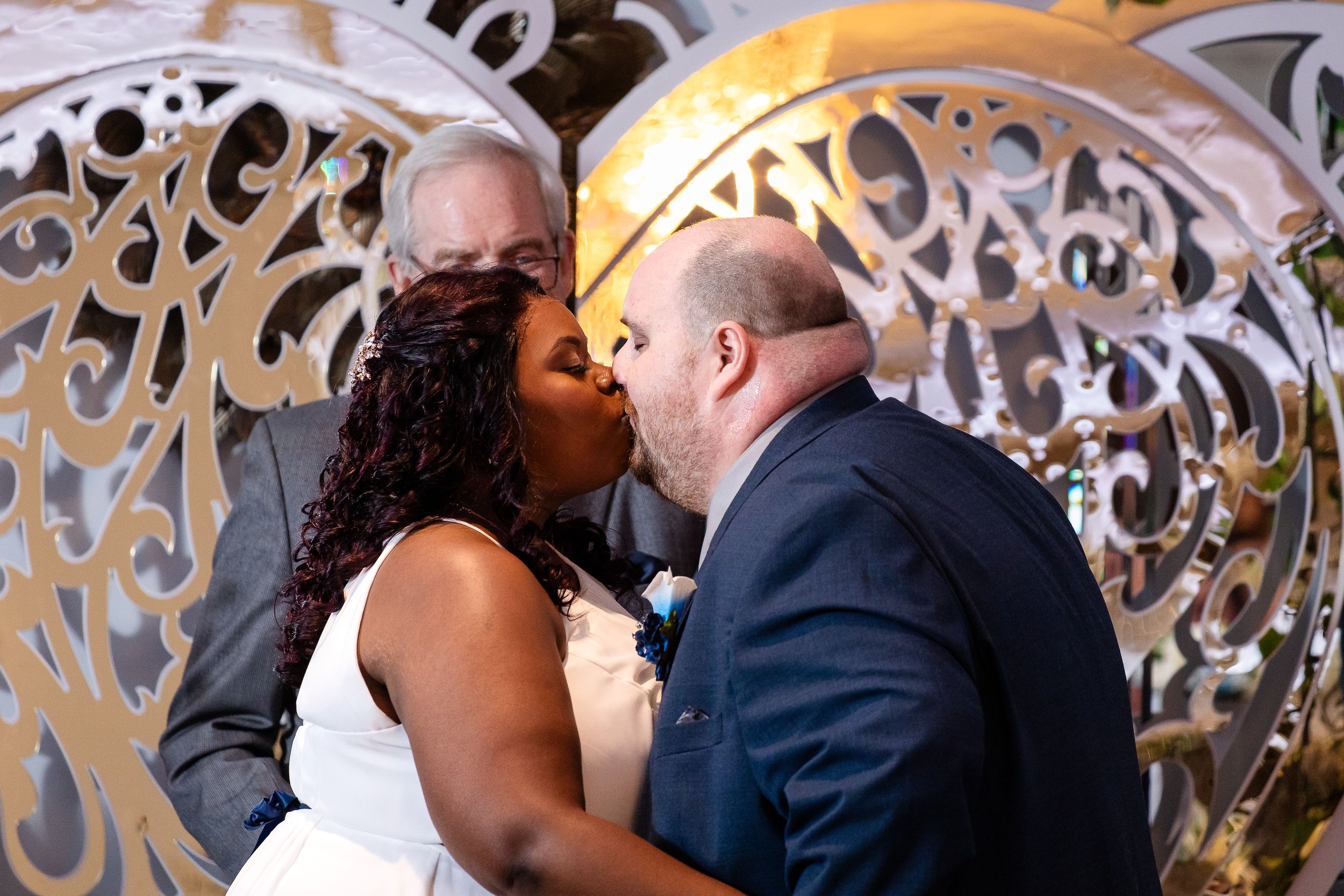 A couple sharing a first kiss at their wedding ceremony before an ornate decorative backdrop.