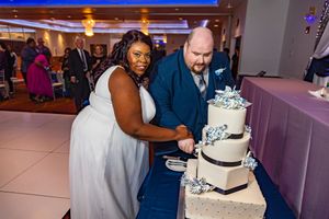 Bride and groom cutting a three-tier white wedding cake with blue floral decorations at their wedding reception.