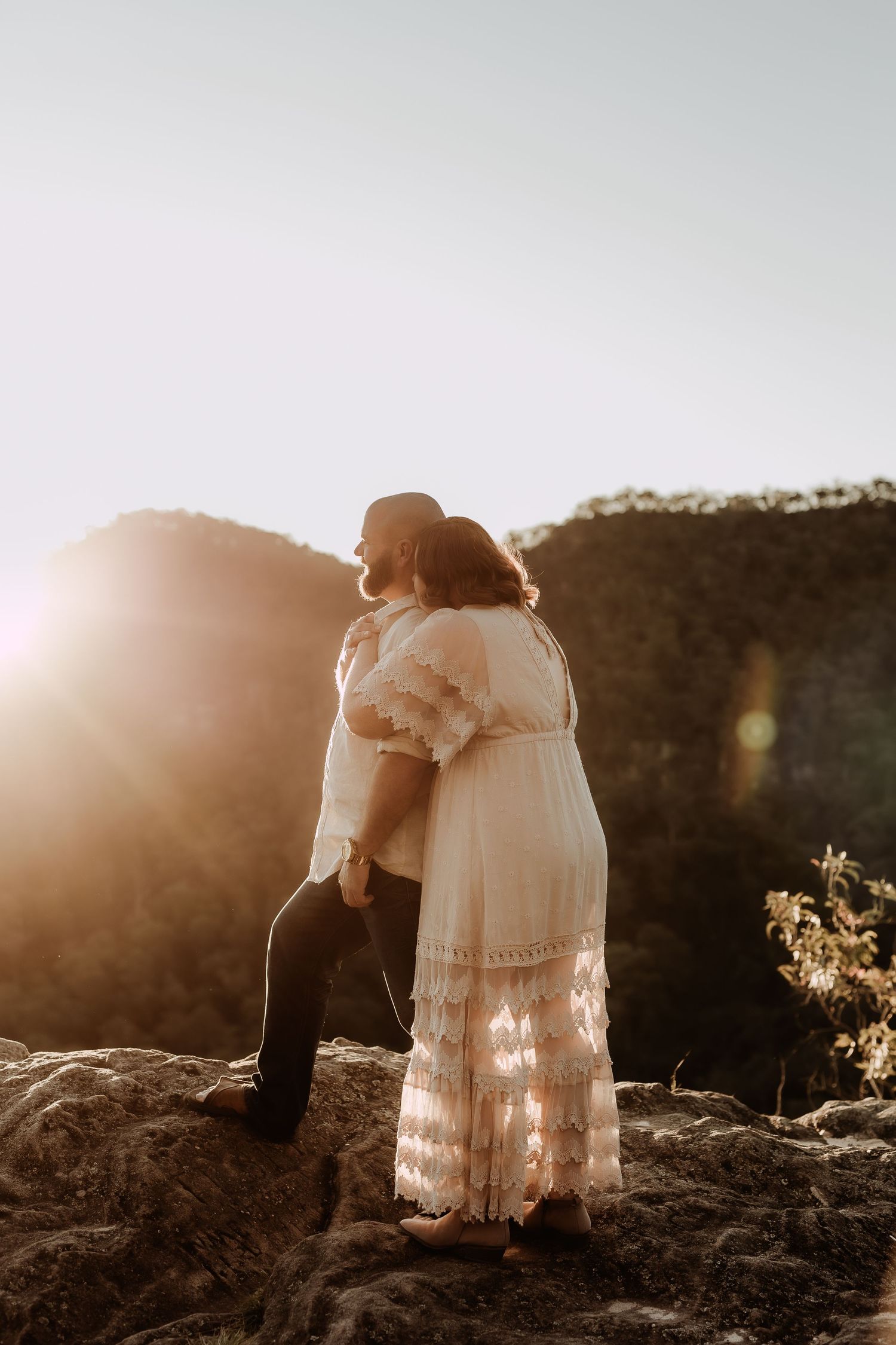 Couple embracing on rocky mountain at golden sunset, woman in white lace boho dress, romantic outdoor engagement photo.