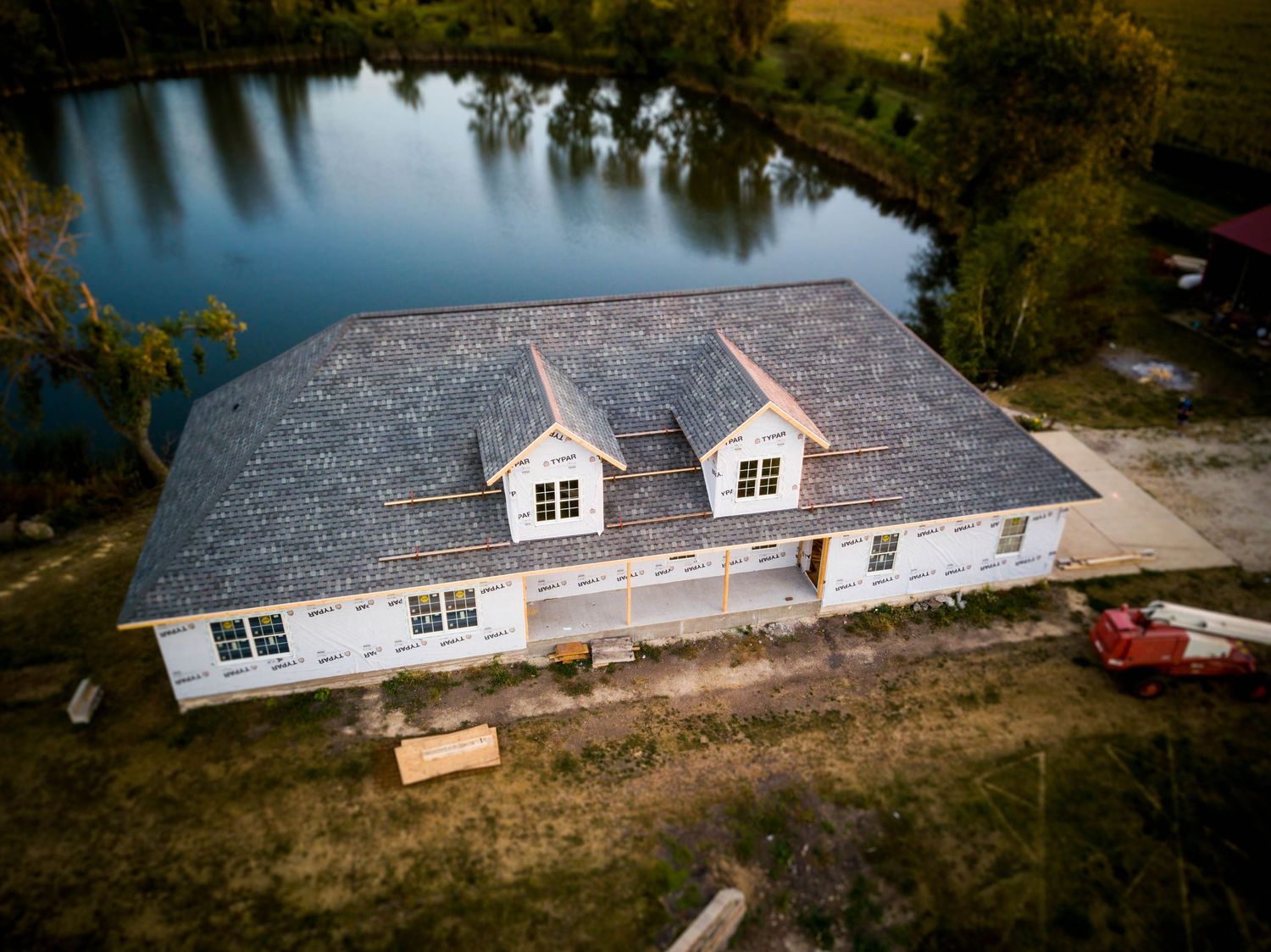 Aerial view of a new construction home with gray shingle roof and dormers beside a serene pond.