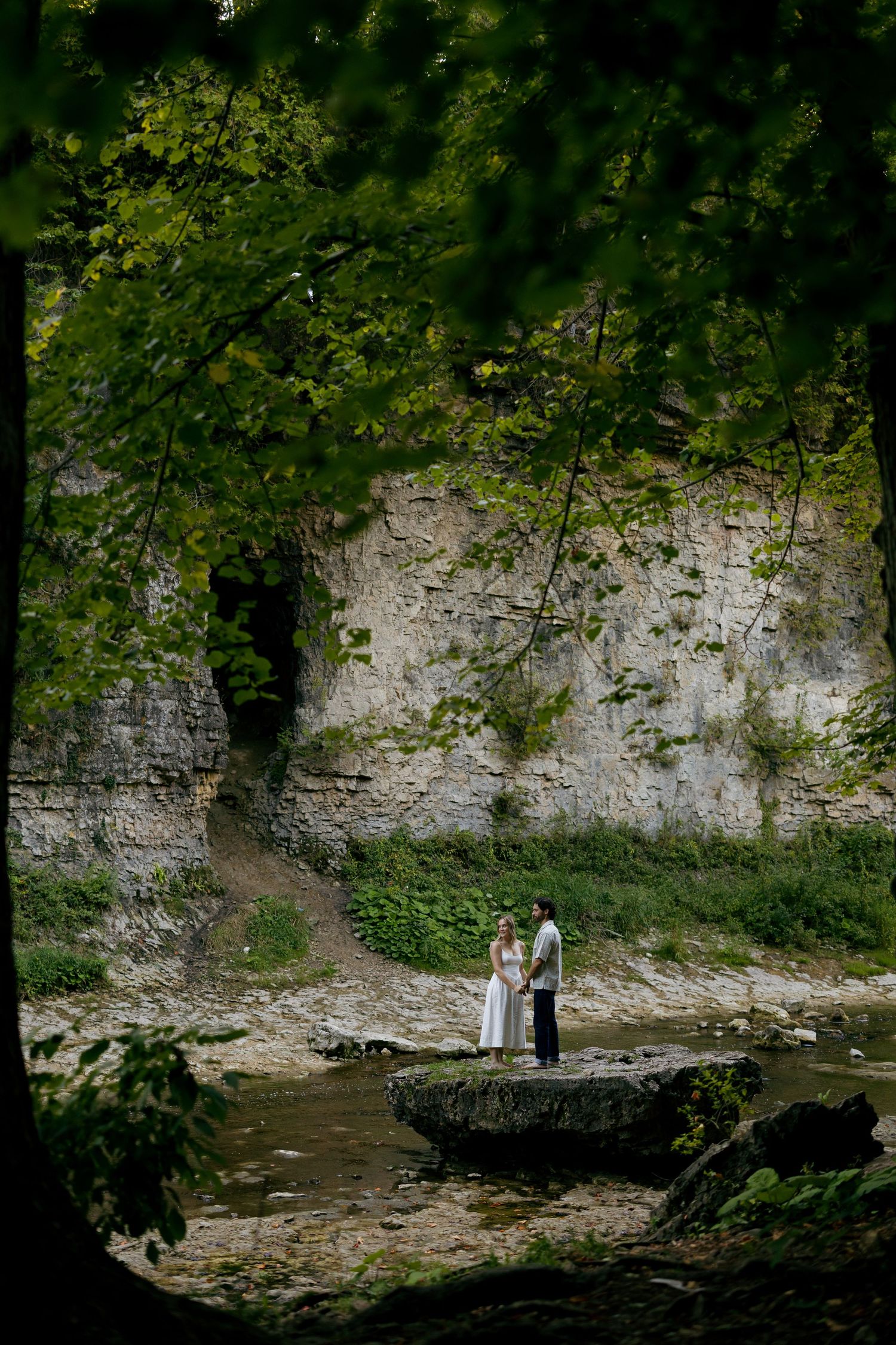 Couple standing on stone platform by creek with white cliff wall and lush green foliage in natural outdoor setting.
