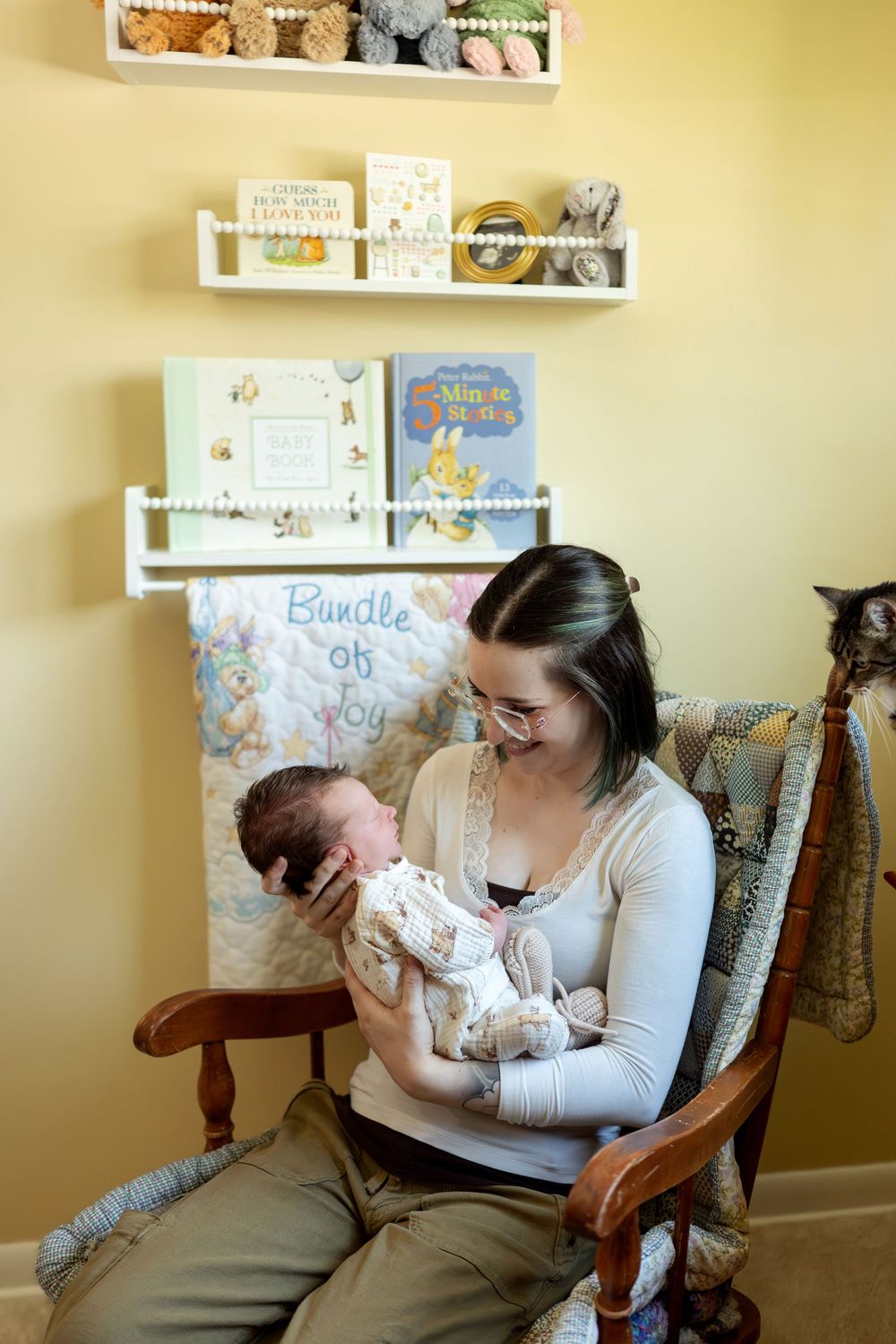 A mother in a white top sits in a wooden rocking chair cradling her newborn in a cozy nursery.