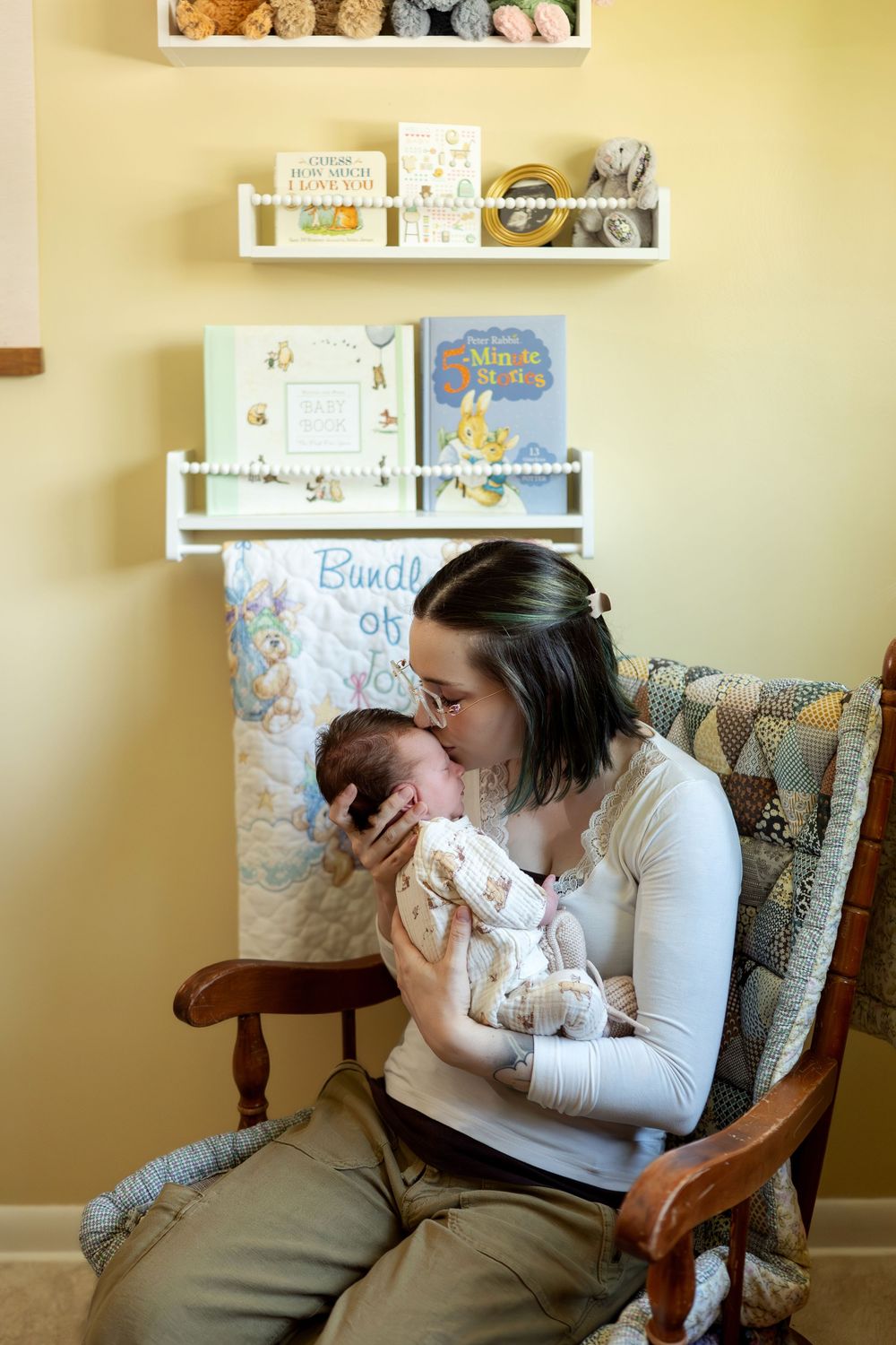 A dark-haired mother gently kisses her sleeping newborn baby while seated in a nursery rocking chair.