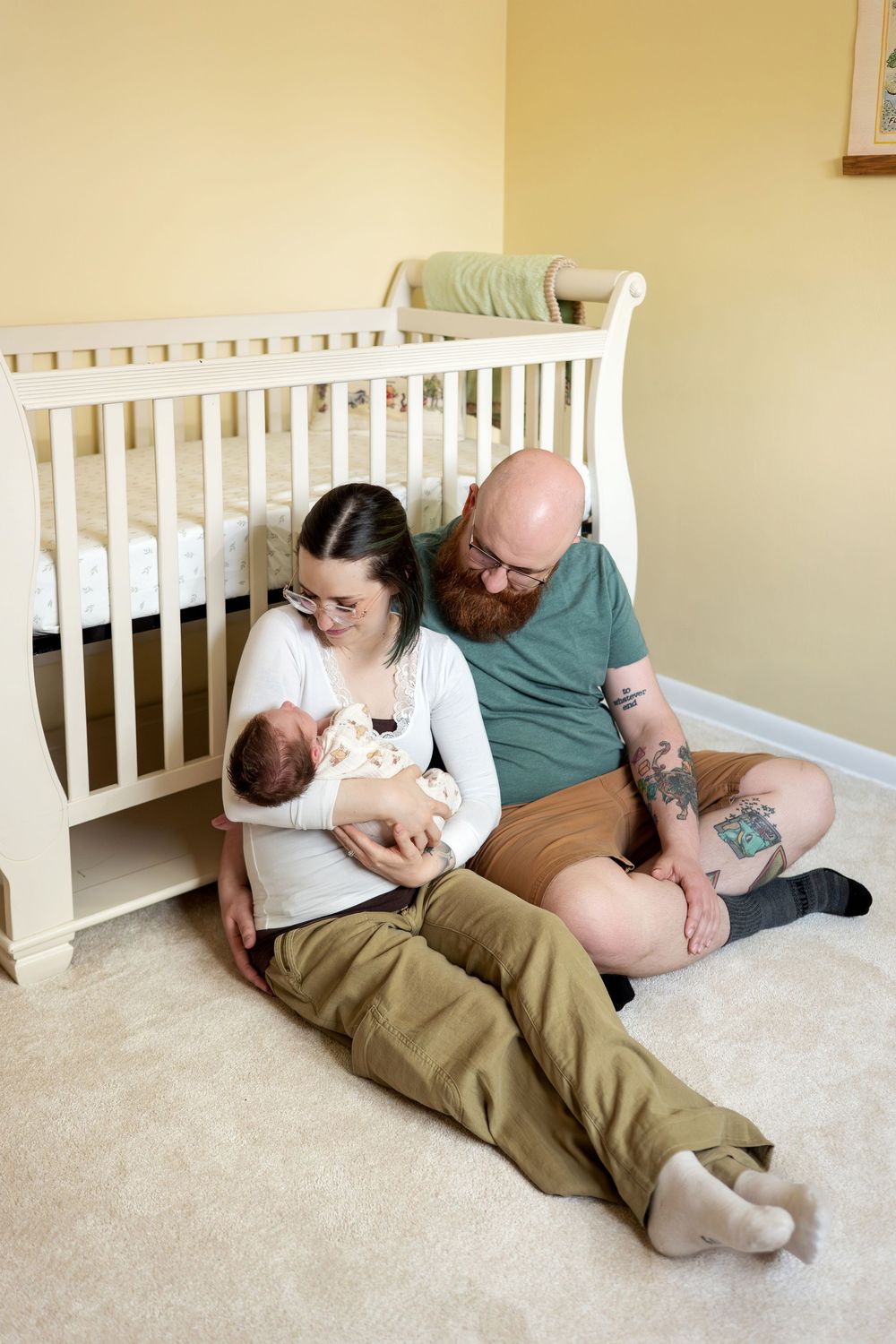 New parents sit on the nursery floor against a white crib, lovingly looking at their newborn baby.