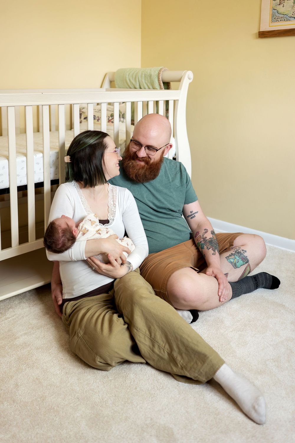 Happy couple sitting on floor in yellow nursery smiling at each other with newborn.