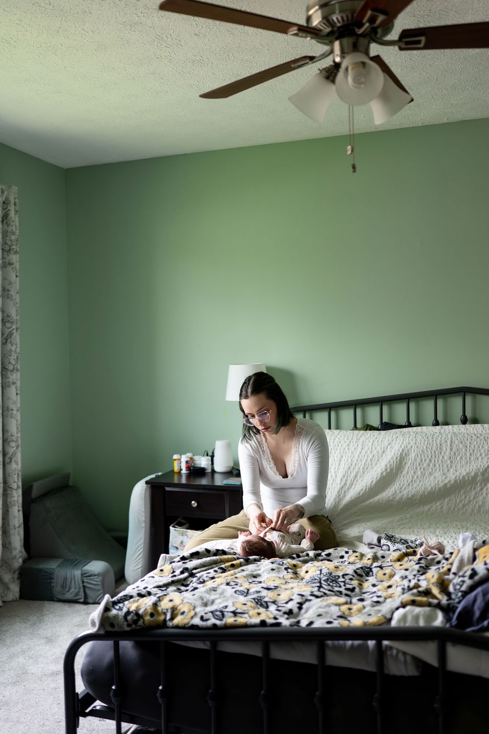 Young mother sits on bed in green bedroom carefully tending to her newborn baby.