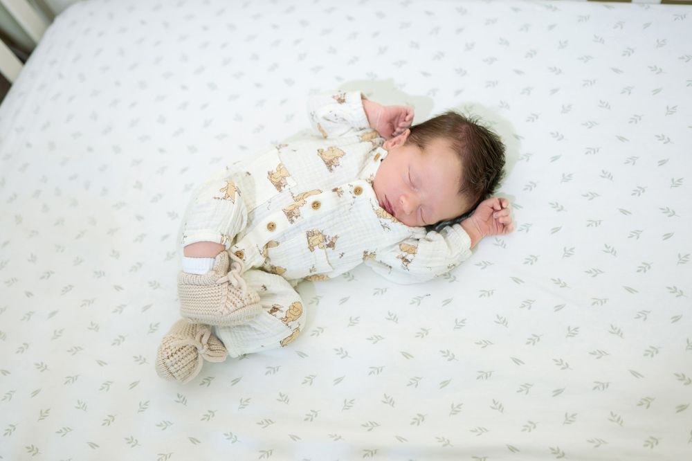 Newborn baby sleeping in white animal print outfit with knit booties on light patterned crib sheet.
