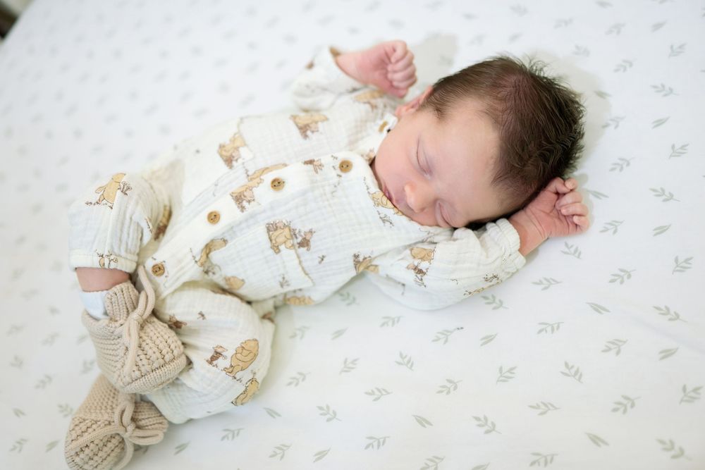 Sleeping newborn in cream animal print outfit with knit booties lying on white leaf print crib sheet.