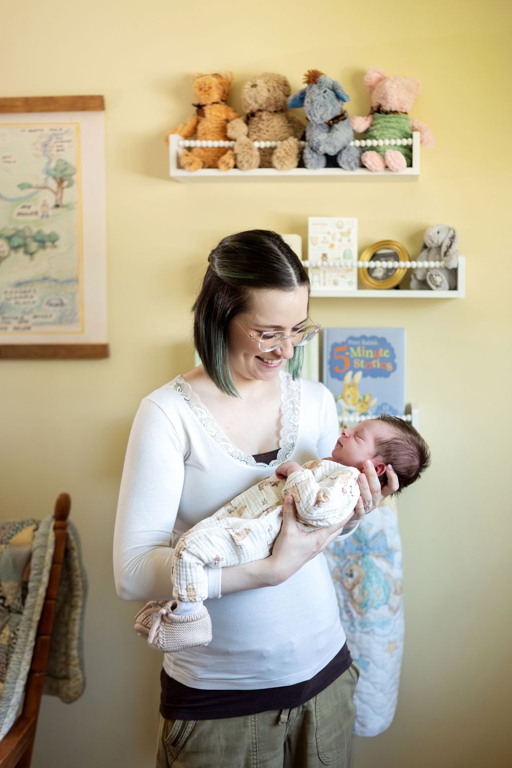 Smiling new mother in white top holding swaddled newborn baby in yellow nursery with stuffed animals.