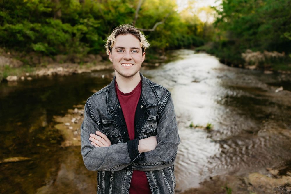 Young man smiling with arms crossed standing by a creek wearing a denim jacket in natural outdoor setting.