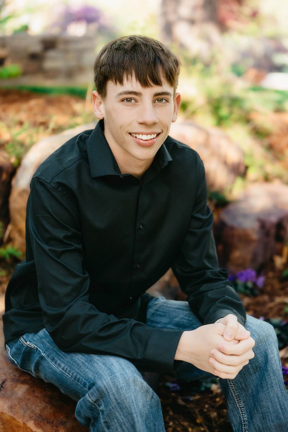 Smiling teenage boy in black shirt and jeans sitting on a rock outdoors in a garden setting.