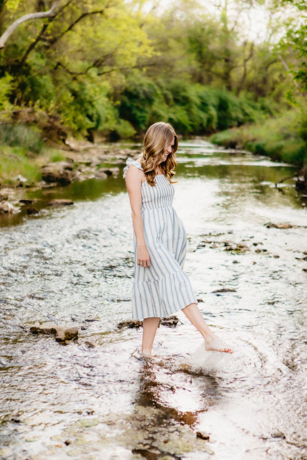 Young woman in striped midi dress wading barefoot in a shallow creek surrounded by lush green trees.