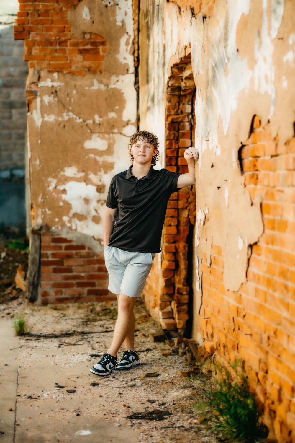 Teen boy in black polo and white shorts leaning against a rustic, weathered brick wall outdoors.