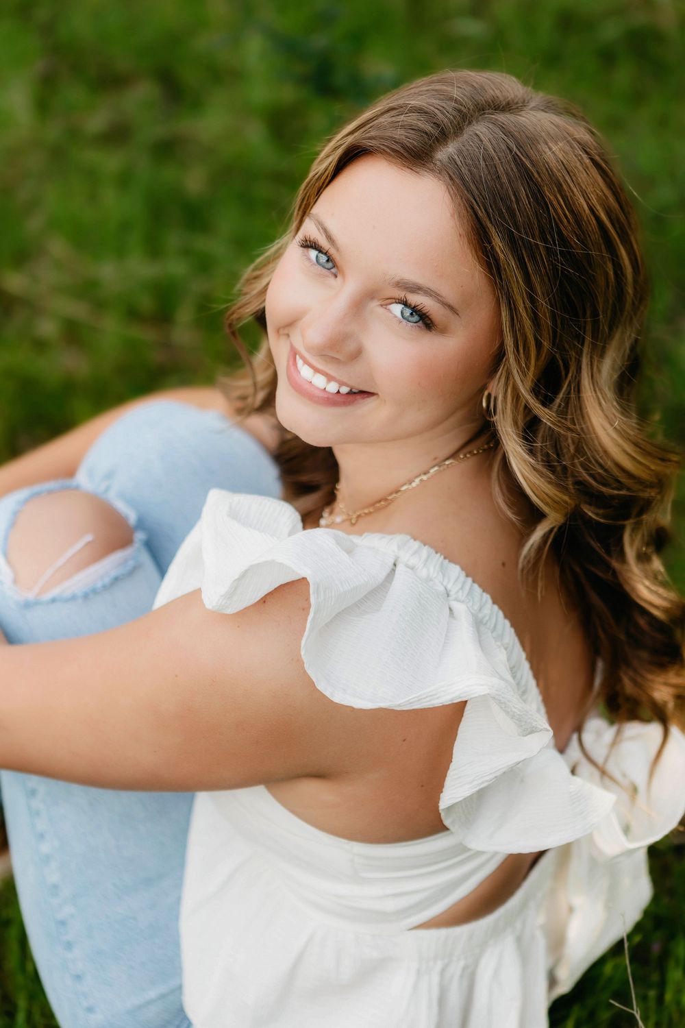 Young woman with blue eyes smiling outdoors, wearing a white off-shoulder top and ripped jeans, sitting on green grass.
