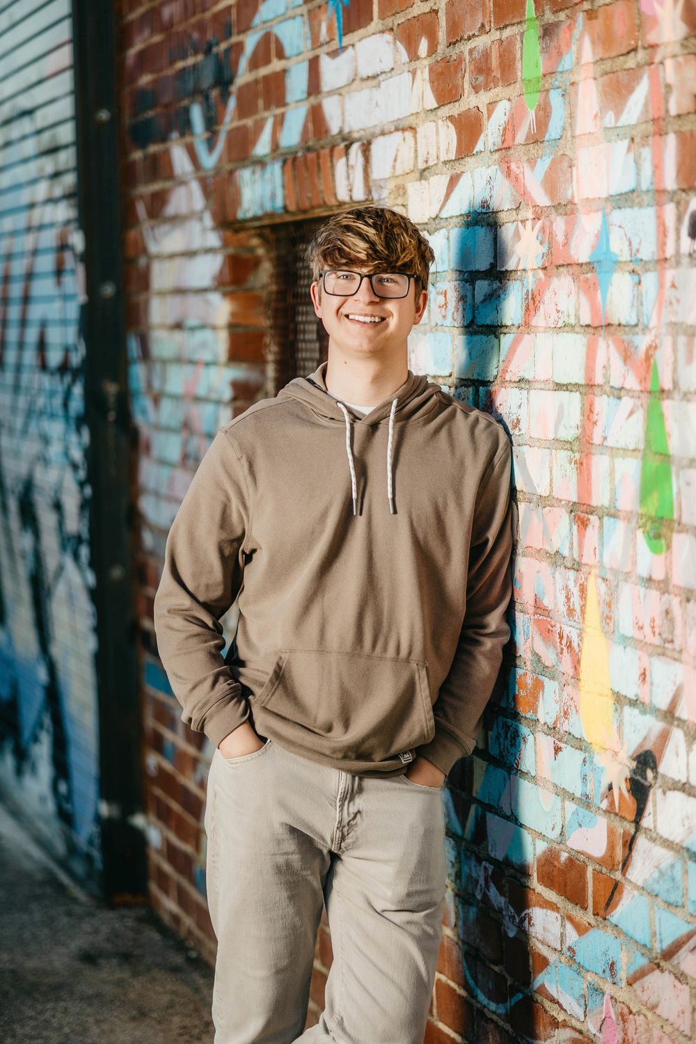 Smiling teen in glasses and brown hoodie leaning against colorful graffiti brick wall.