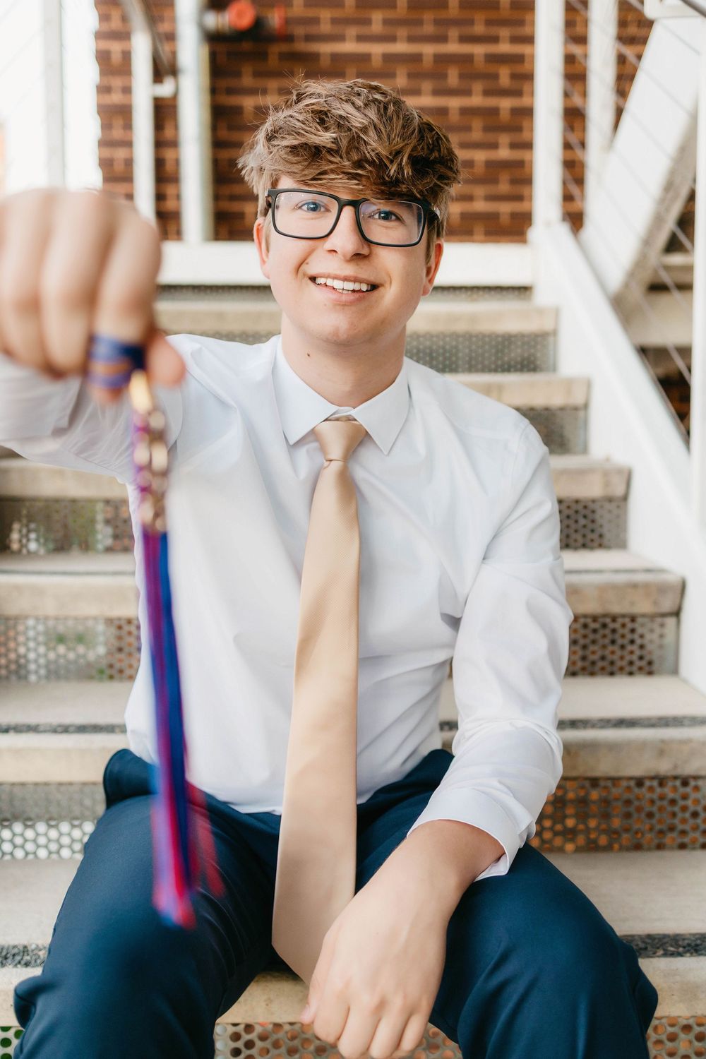Smiling teen boy in white shirt and tan tie sits on steps holding a medal toward camera, wearing glasses.