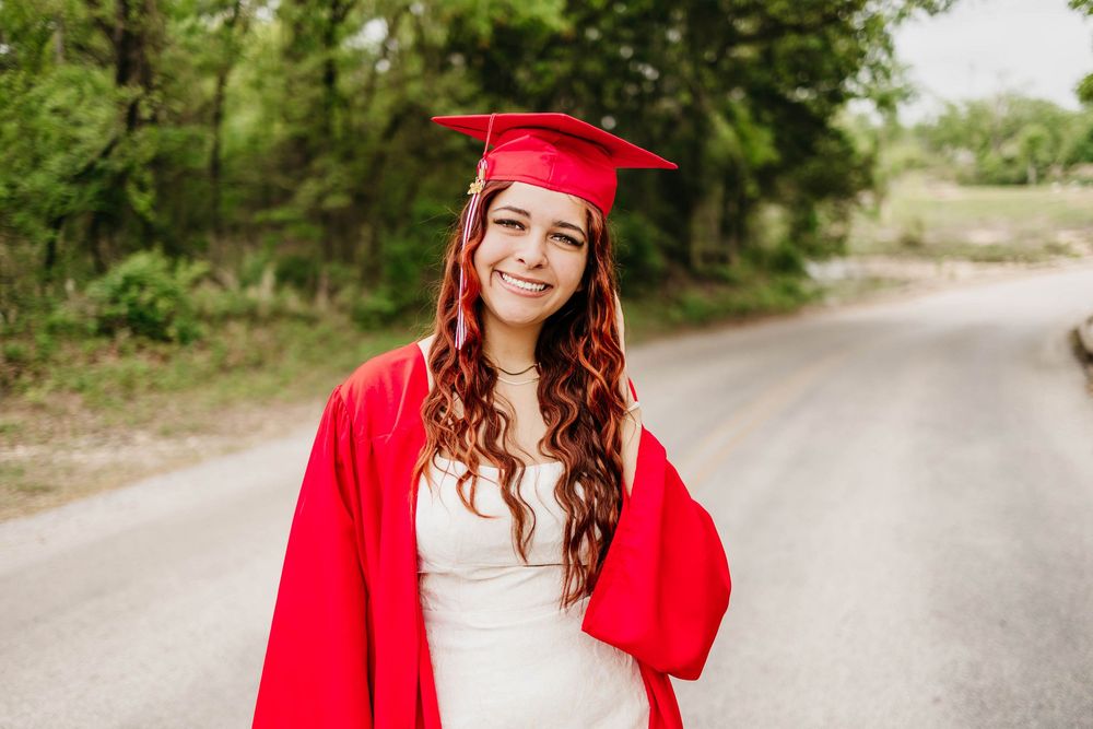 Smiling young woman in red graduation cap and gown standing on a tree-lined road for senior portrait.