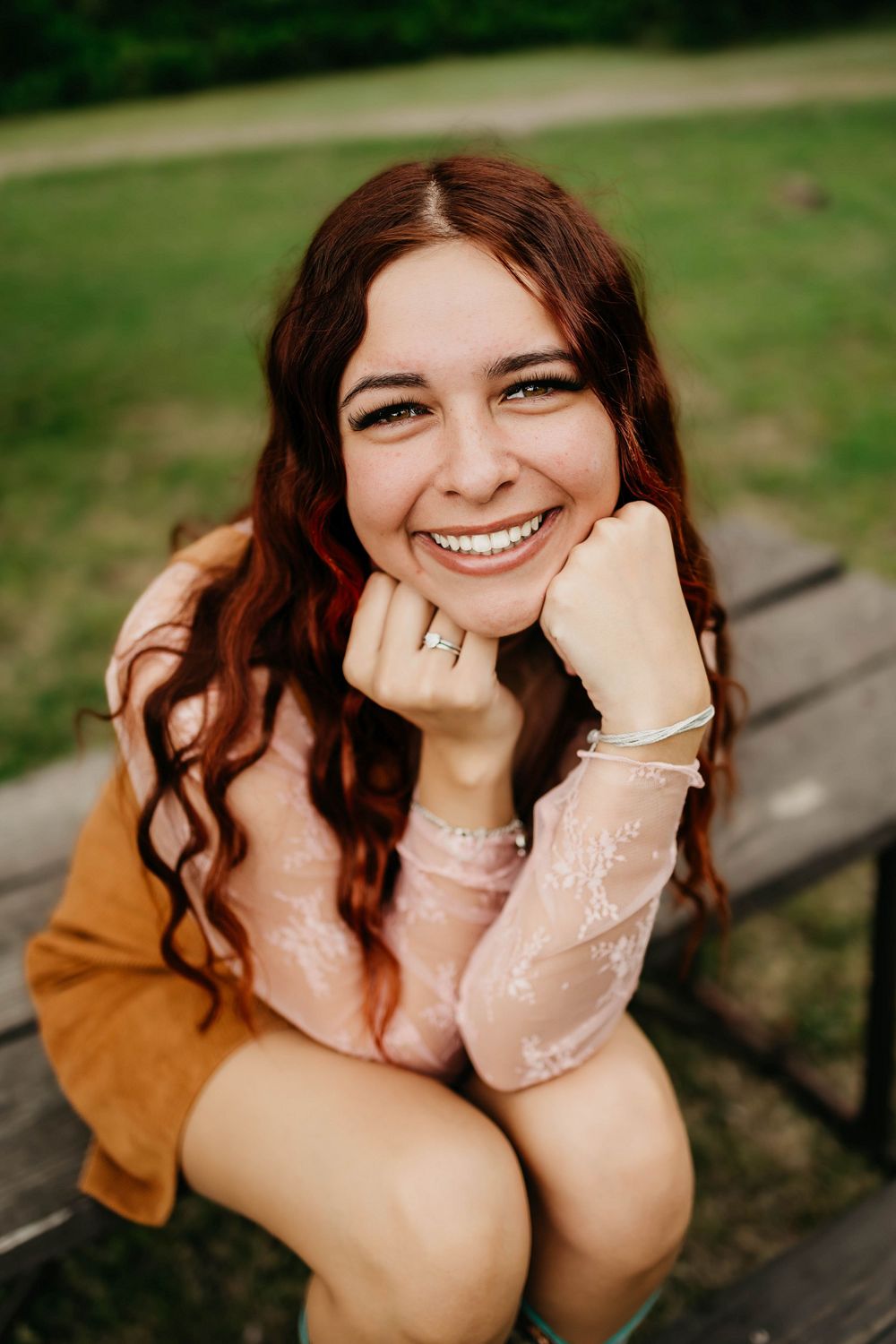 Smiling young woman with long red wavy hair sitting on a bench outdoors, wearing a pink lace top and brown cardigan.