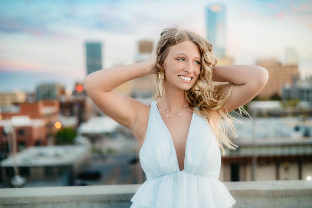 Woman in white dress smiling on rooftop with city skyline in background during golden hour.