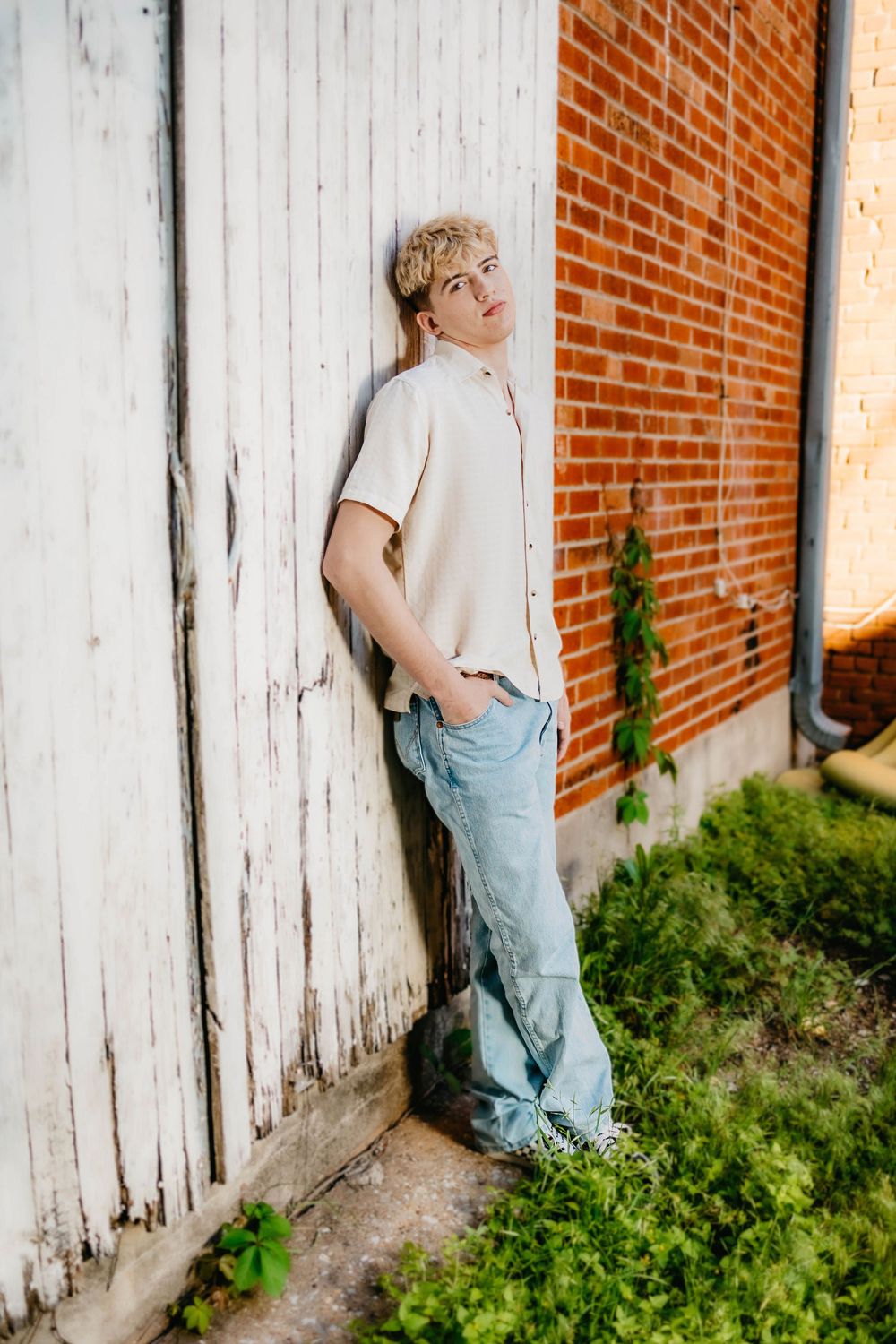 Young man with blonde hair leans against white wooden wall near brick building wearing light shirt and jeans.