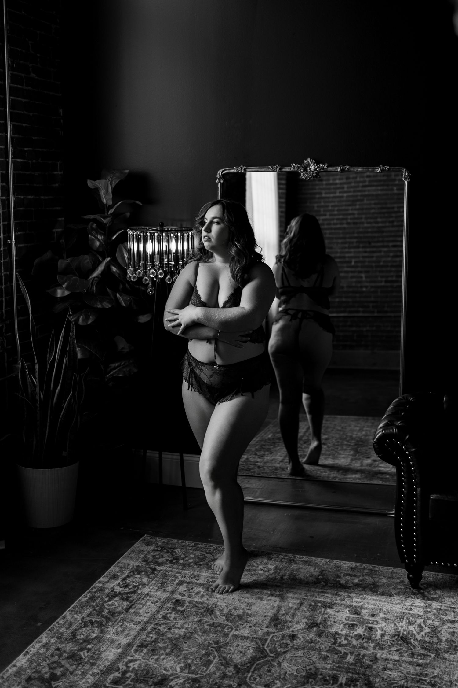Woman in red lace panties posing near window with brick wall and mirror reflection in moody boudoir setting.