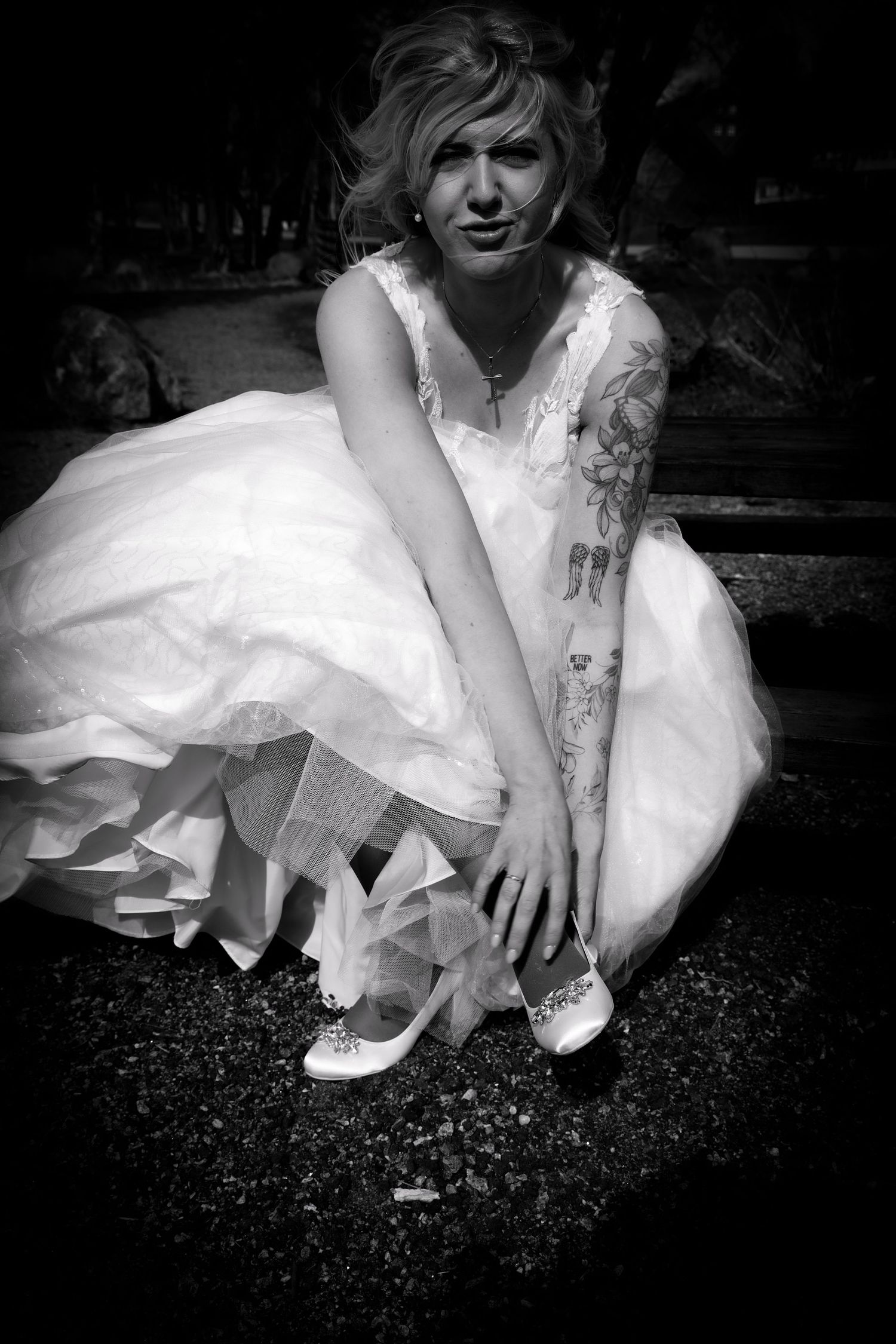 Tattooed bride in white ball gown crouching to adjust her heels in a black and white portrait.