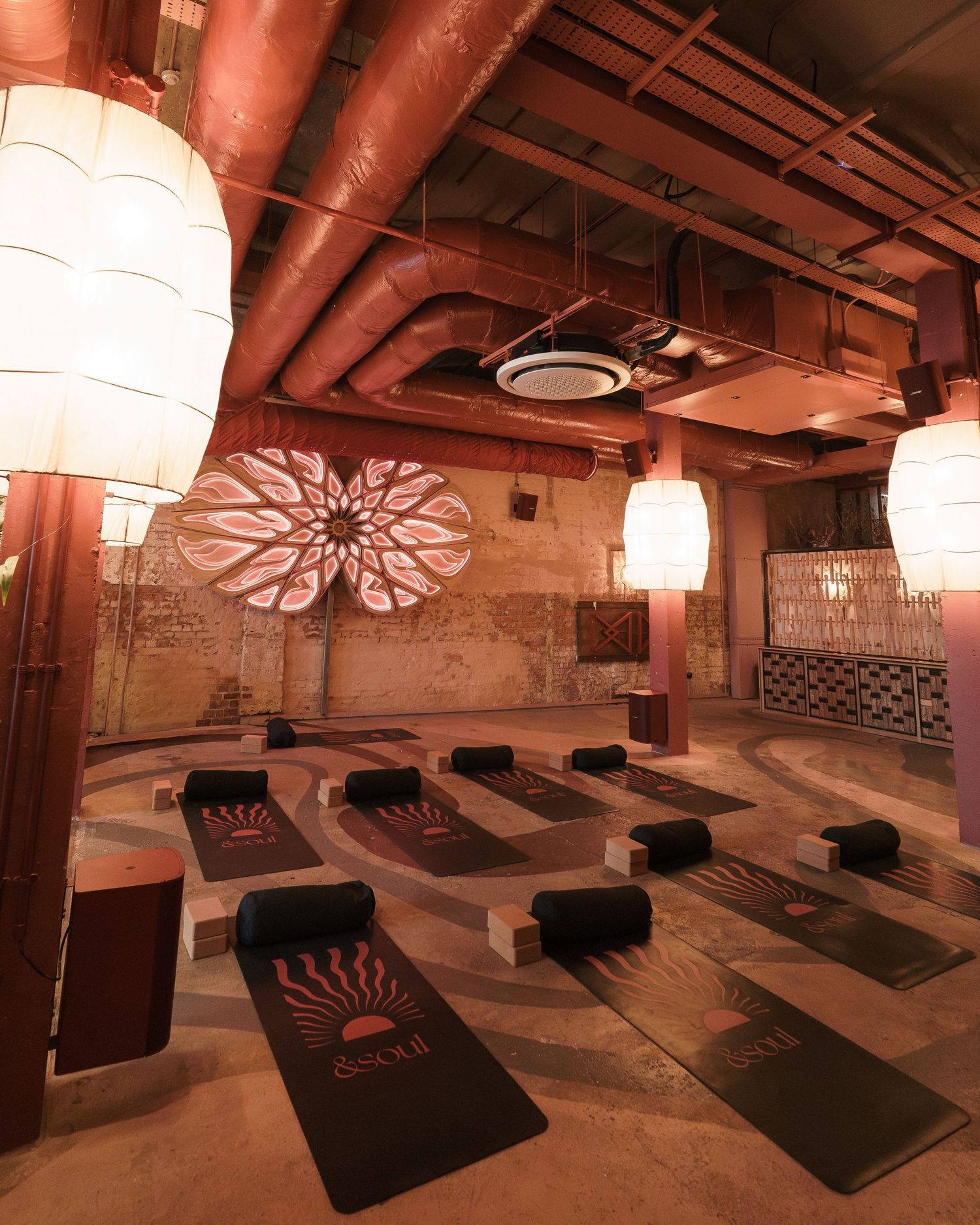 Serene yoga studio with warm lighting, decorative rose window mural, aligned dark mats, and hanging lanterns.