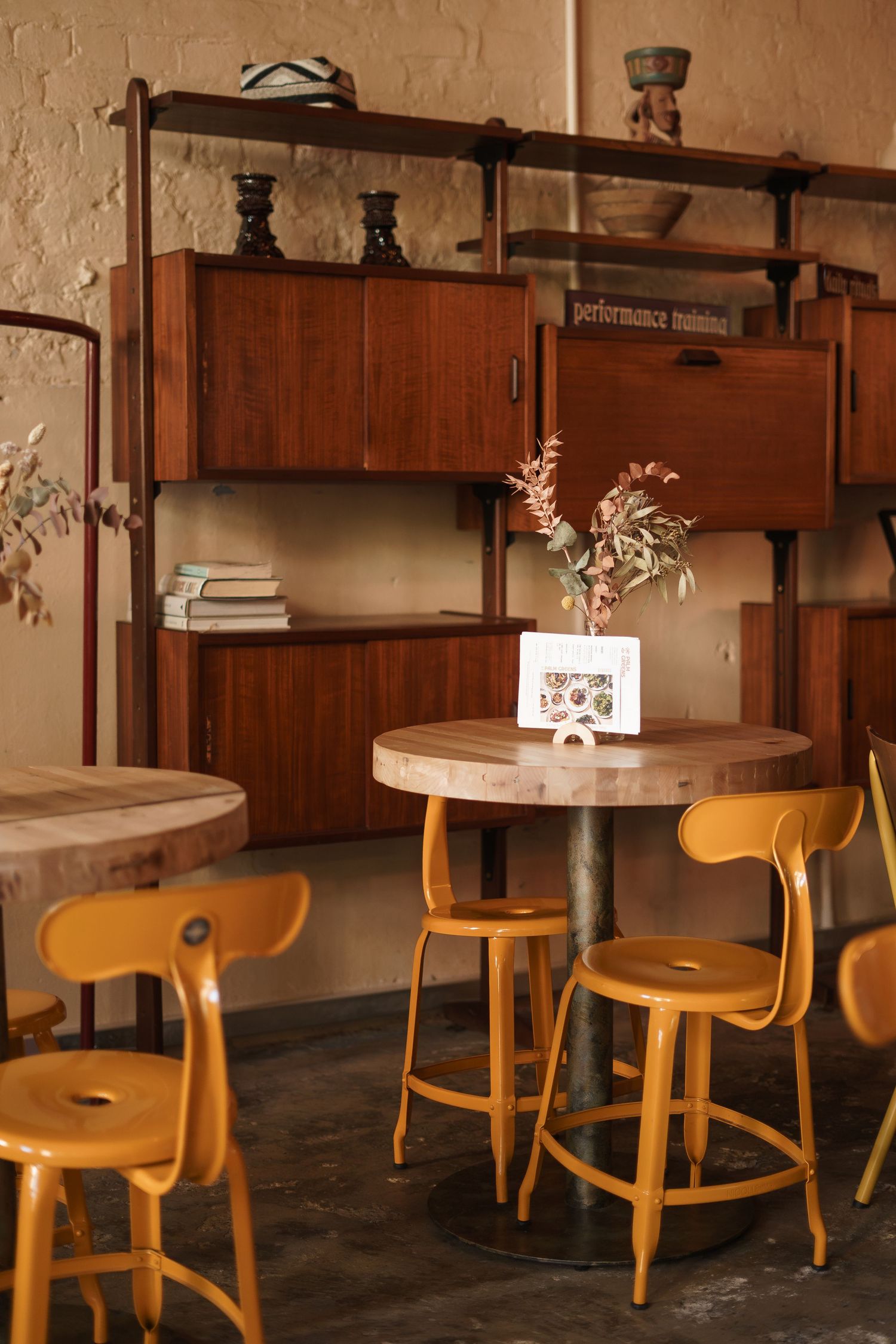Cozy cafe interior with yellow stools, round wooden table, and mid-century modern shelving unit against a warm beige wall.