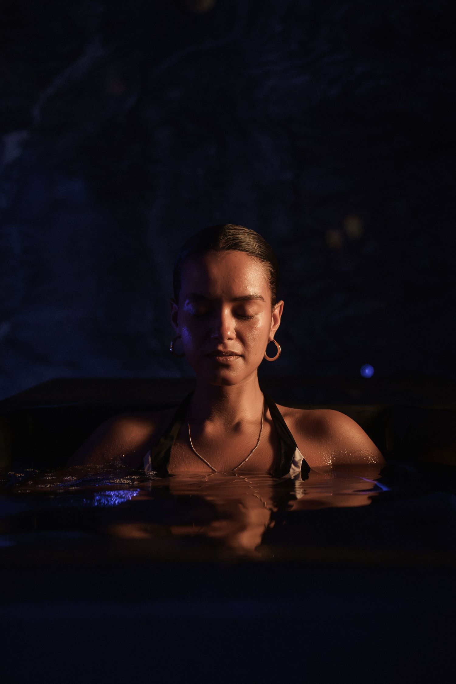 Woman with closed eyes relaxing in a dark pool at night, wearing gold hoop earrings and a necklace.