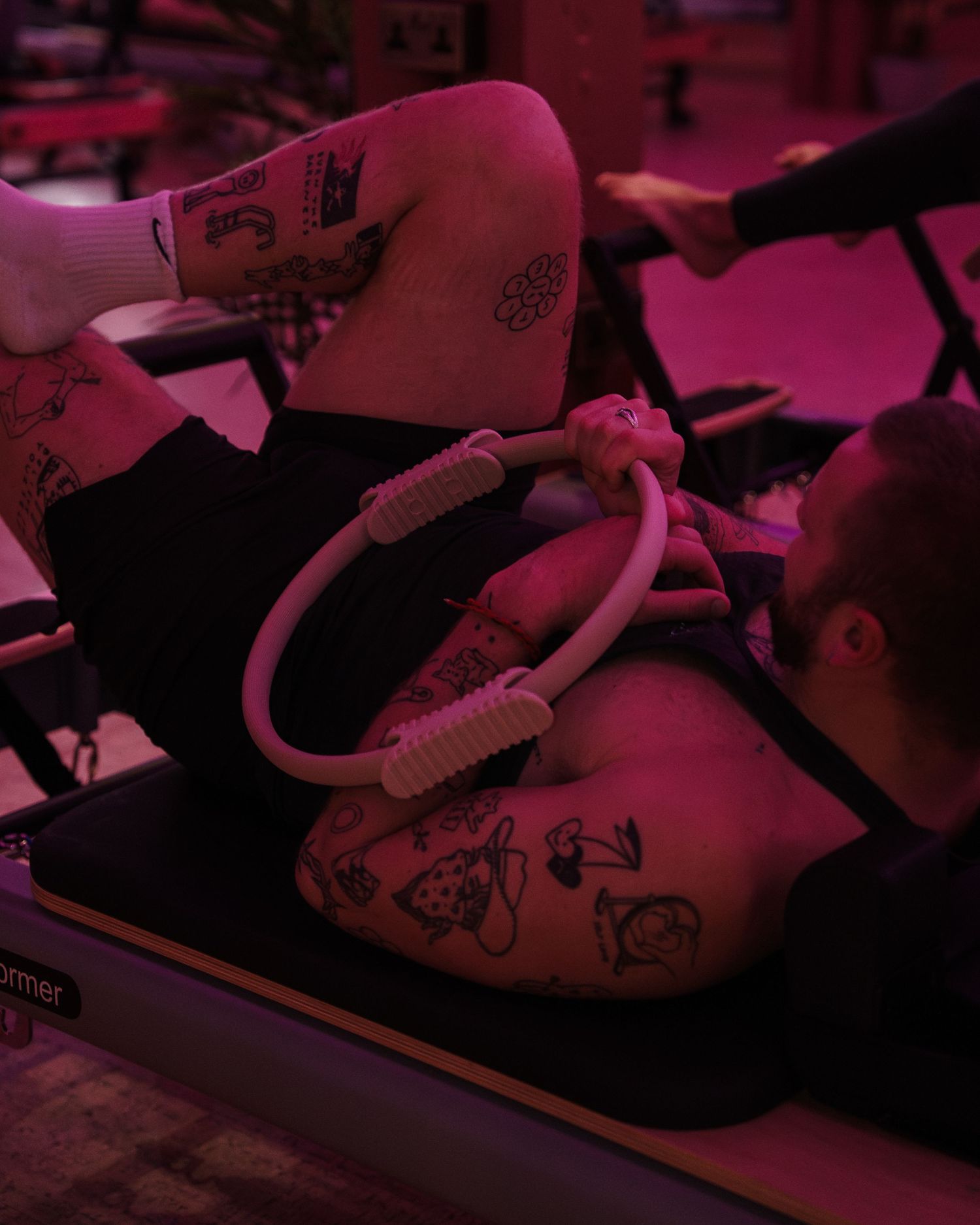 Tattooed person using a pilates ring on a reformer machine under pink studio lighting.