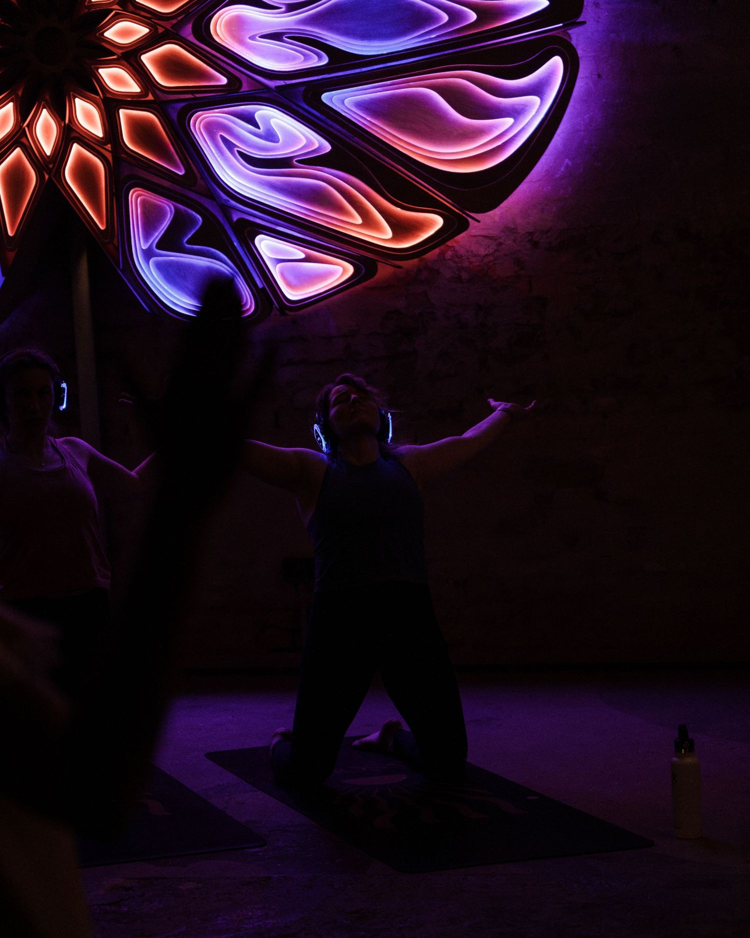 Person doing yoga beneath a glowing neon mandala light installation in a dark purple-lit room.
