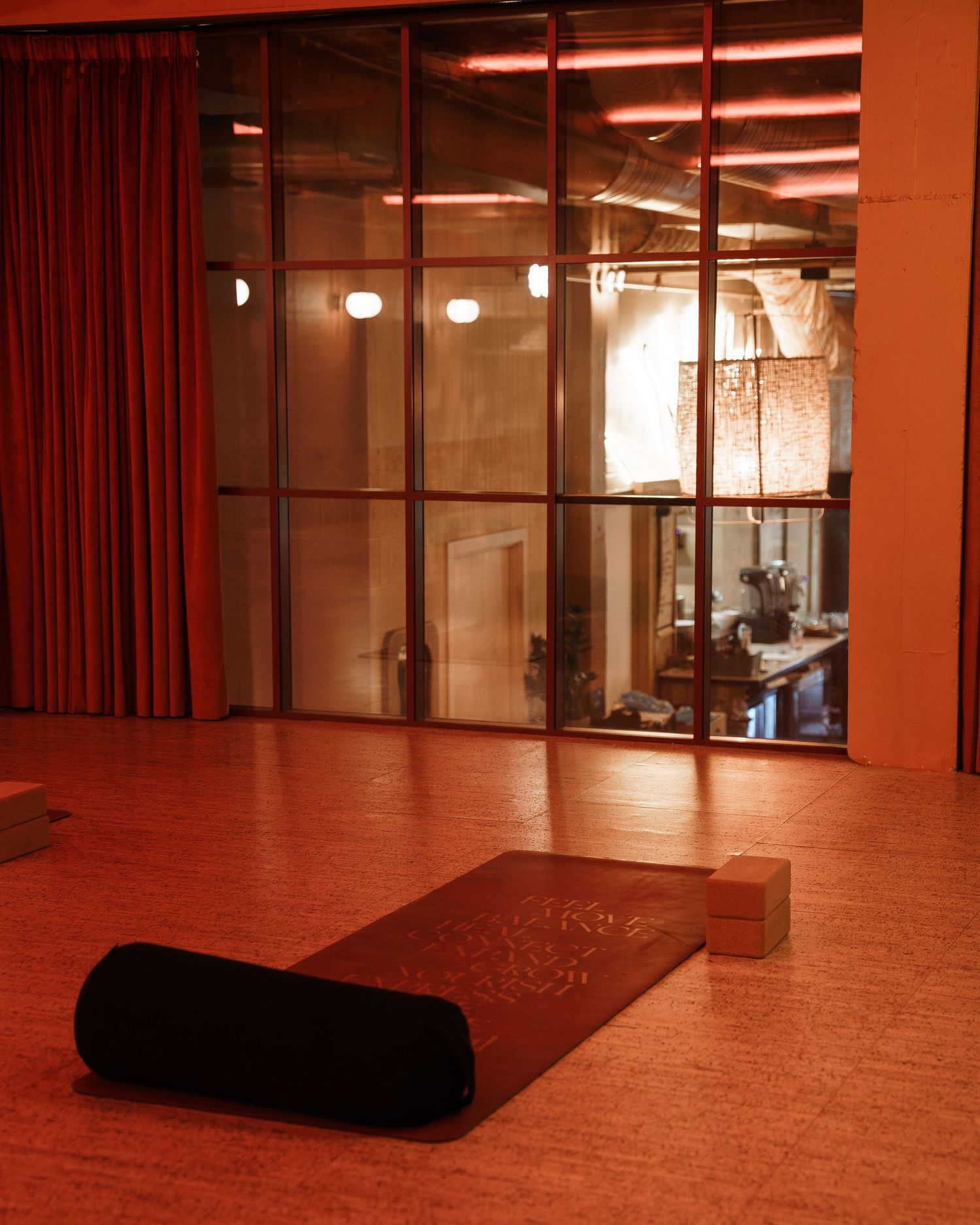 A yoga studio with cork floors, a bolster mat, and glass partition walls bathed in warm red ambient lighting.