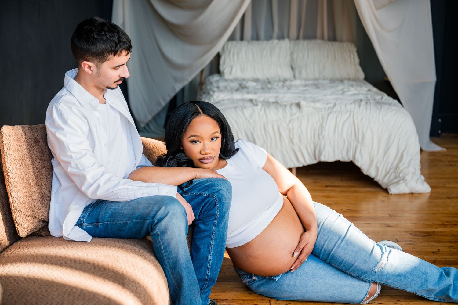 Couple sharing a tender moment, pregnant woman in jeans resting against partner near a bed in Hartford, CT photo studio.