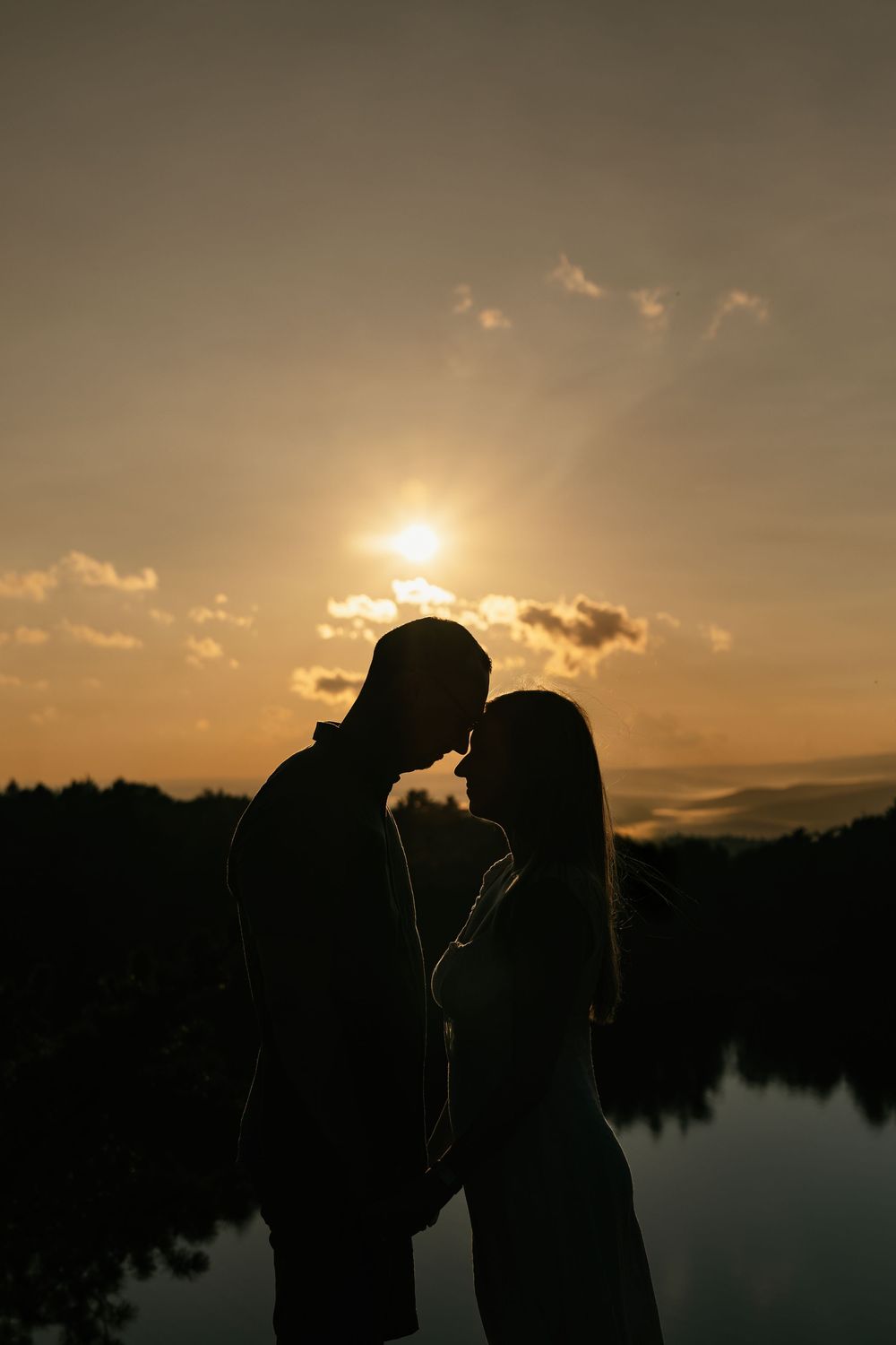 Romantic couple silhouette facing each other against a golden sunset sky reflected on a calm lake.