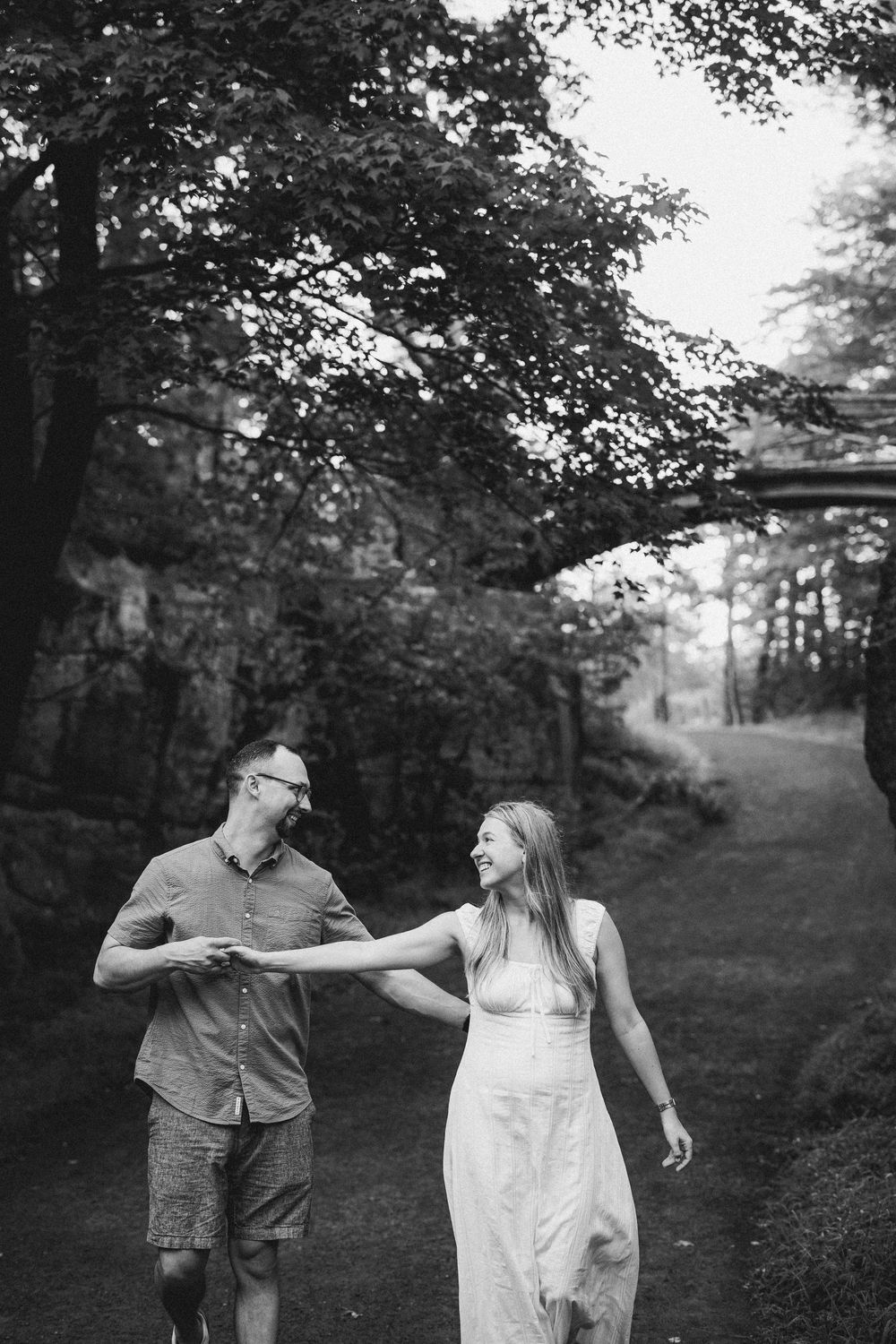 A couple holds hands and smiles while walking along a tree-lined path in black and white.