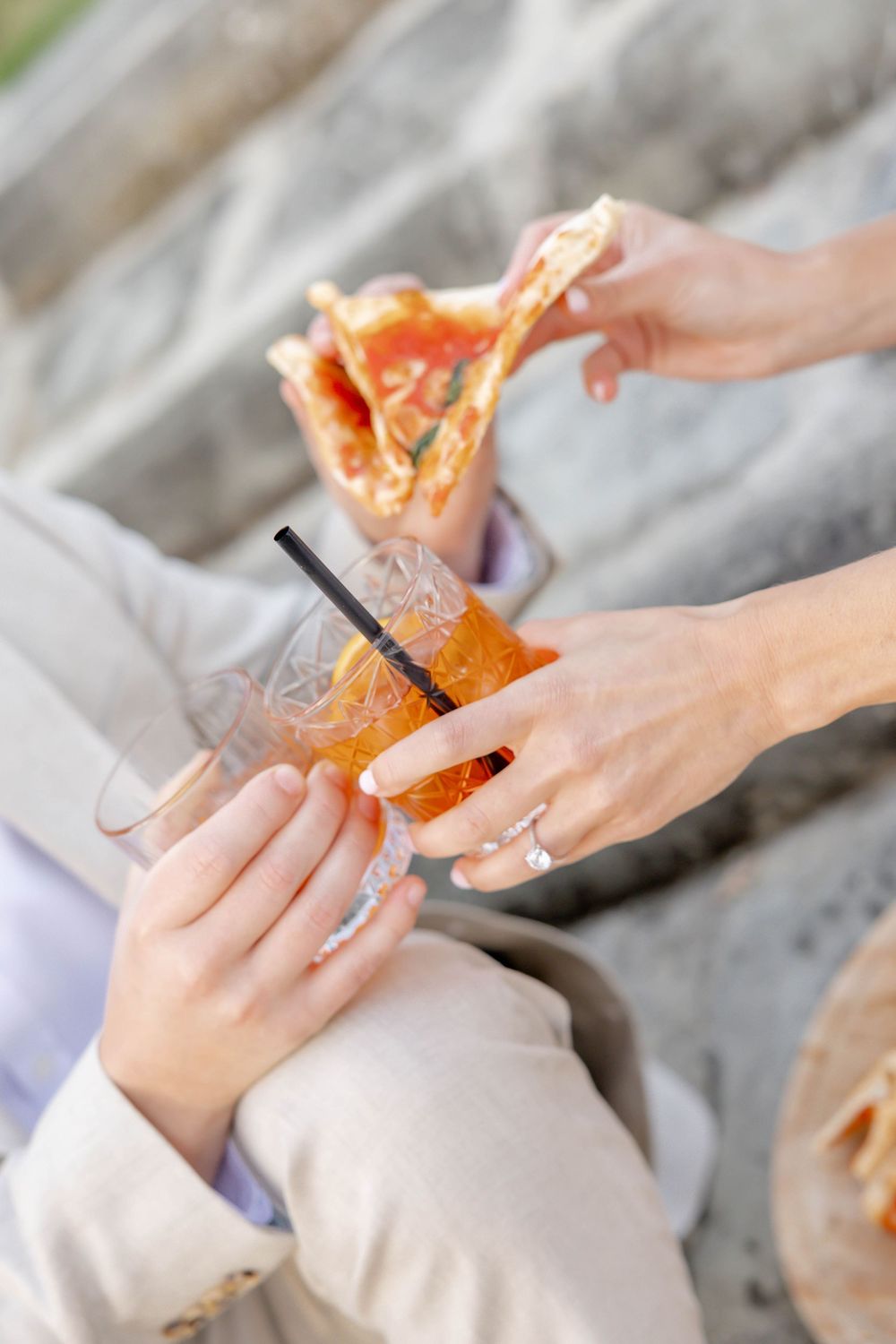 Two people clinking glasses of Aperol Spritz while sharing a slice of pizza outdoors.