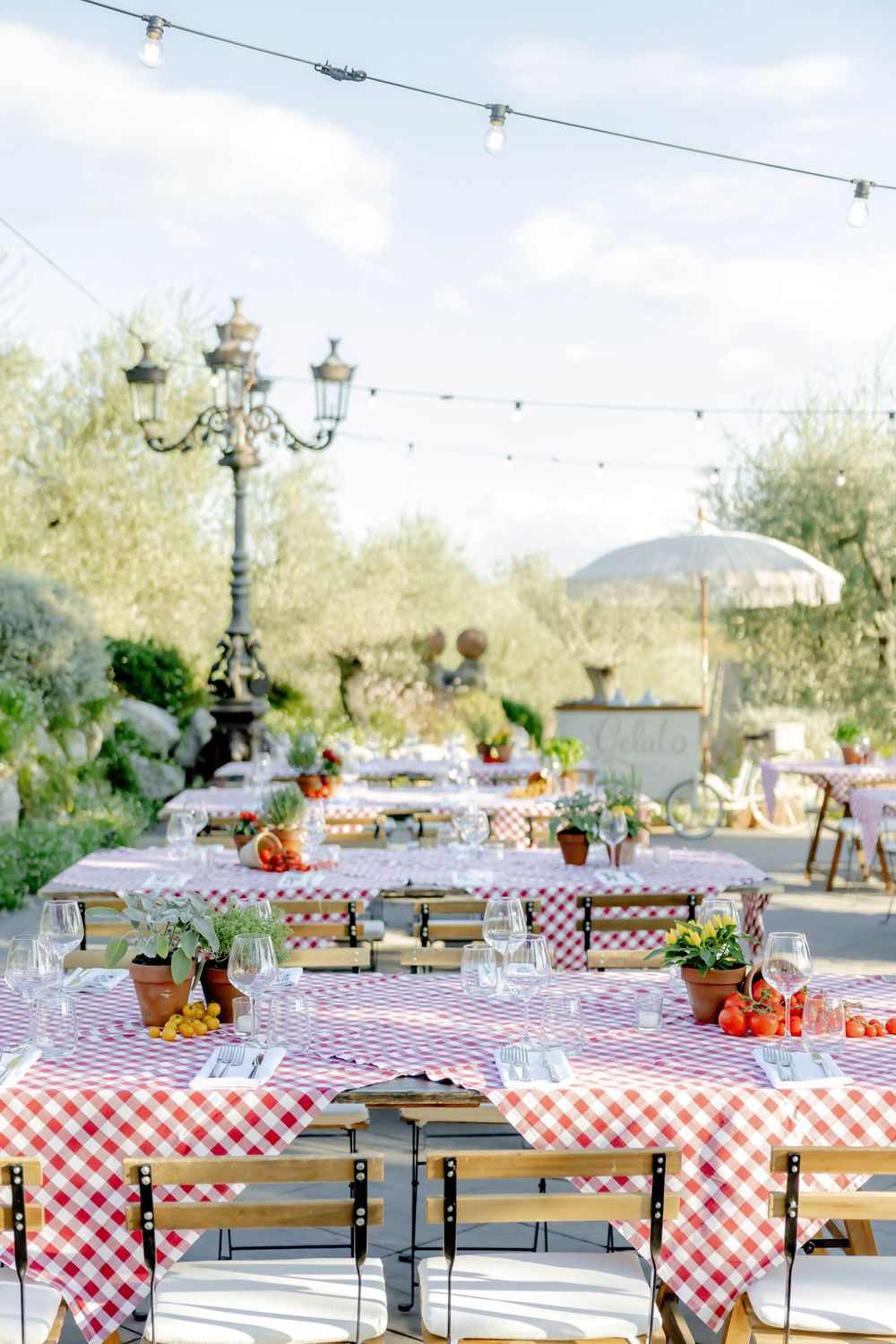 Outdoor dining tables with red gingham tablecloths, wine glasses, and fresh flowers under string lights in a garden setting.