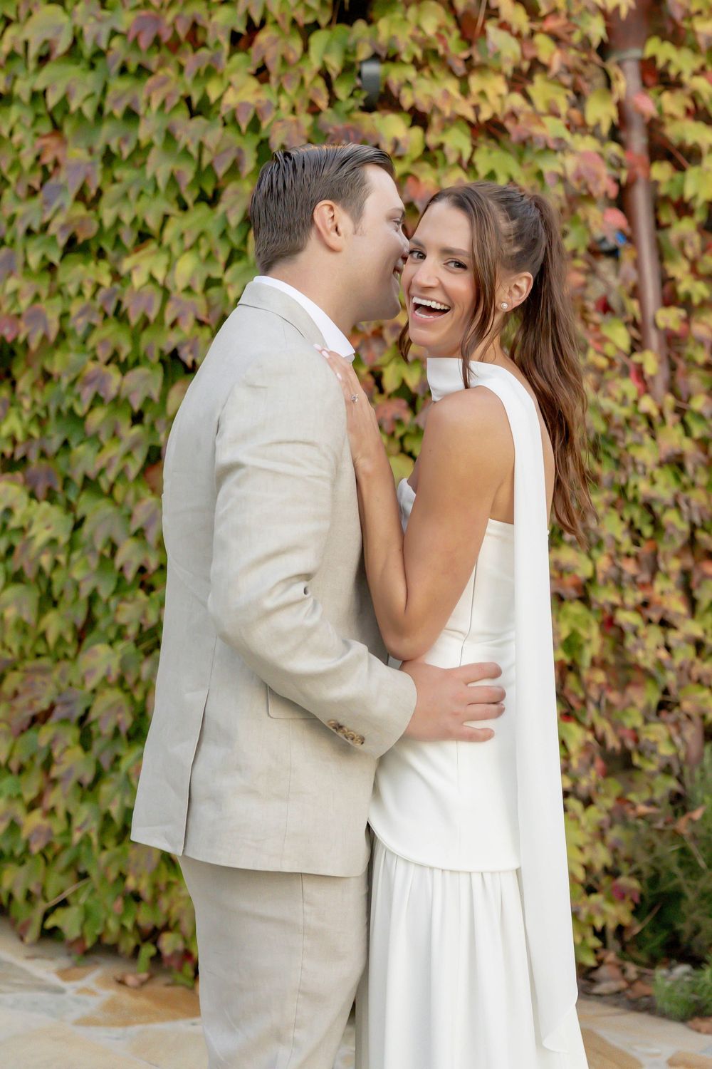 A smiling bride in a white halter dress embraces a groom in a beige suit before an ivy-covered wall.