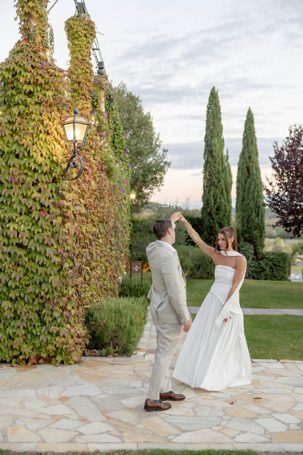 Bride and groom dancing on stone patio surrounded by ivy-covered walls and cypress trees at sunset.