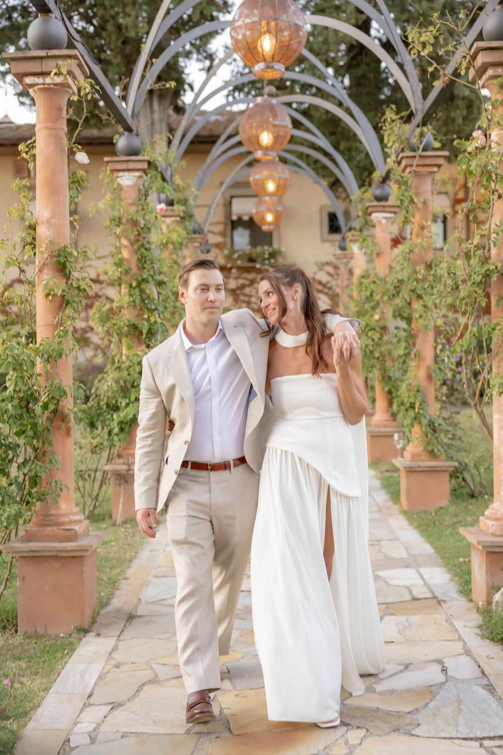 A couple in wedding attire walks under an arched pergola with hanging lights and lush greenery at a romantic venue.