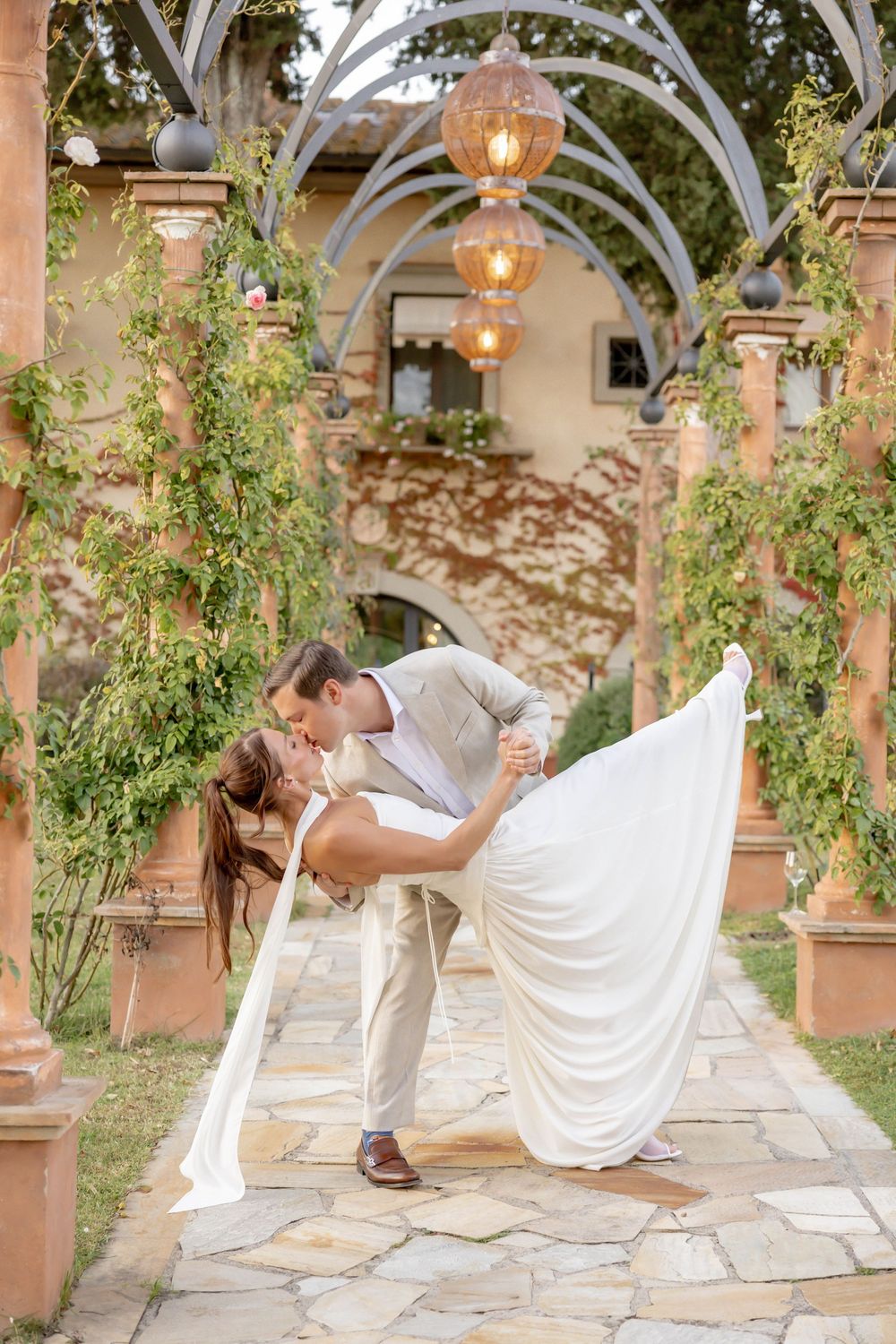 Bride and groom share a romantic dip kiss under an arched garden pergola with hanging lanterns at their wedding.