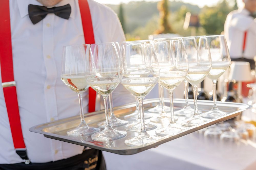 Waiter in red suspenders and bow tie serving white wine glasses on a silver tray at an outdoor event.