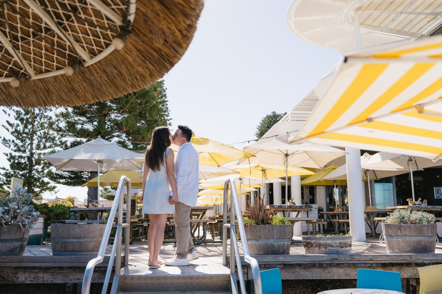 Couple kissing on steps of a beachside cafe with yellow striped umbrellas and outdoor seating on a sunny day.