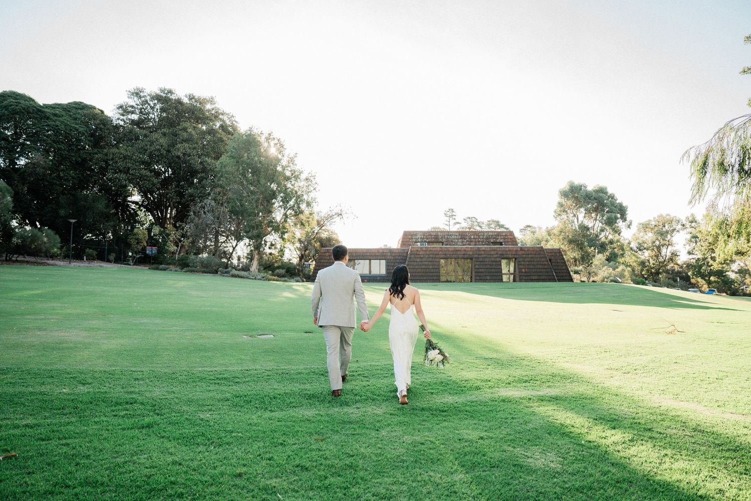 Bride and groom walking hand-in-hand across lush green lawn toward rustic building surrounded by trees.