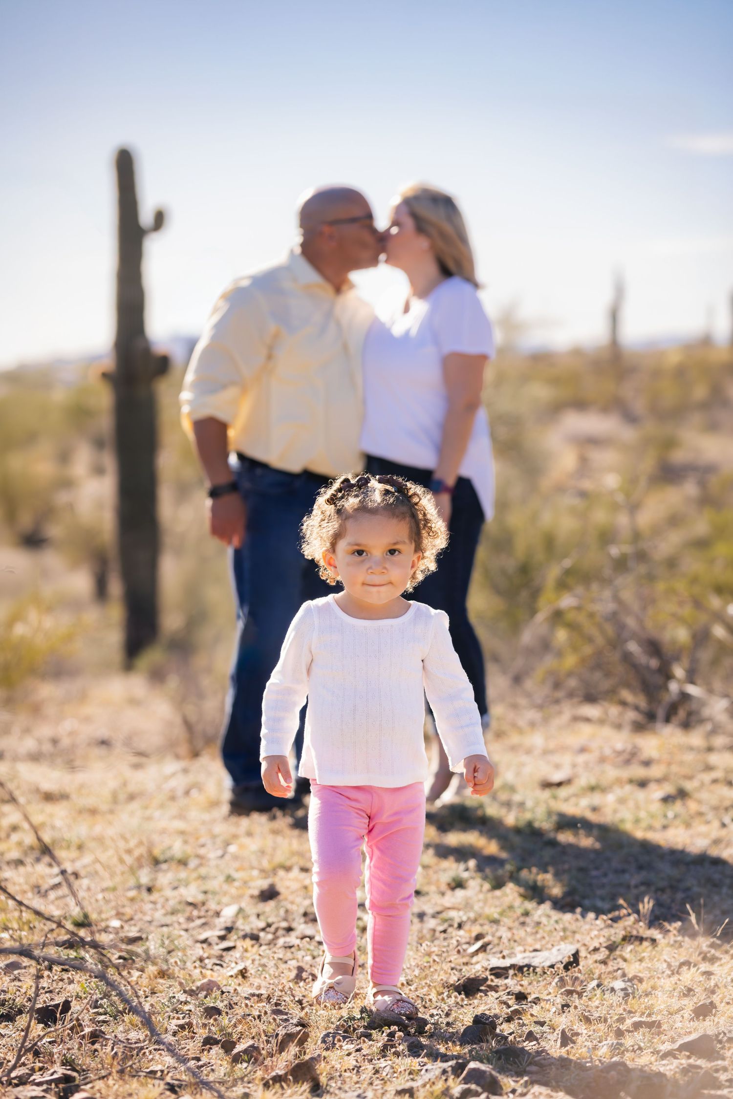 A young toddler girl stands in front of her parents kissing in a desert landscape with saguaro cacti in Arizona.