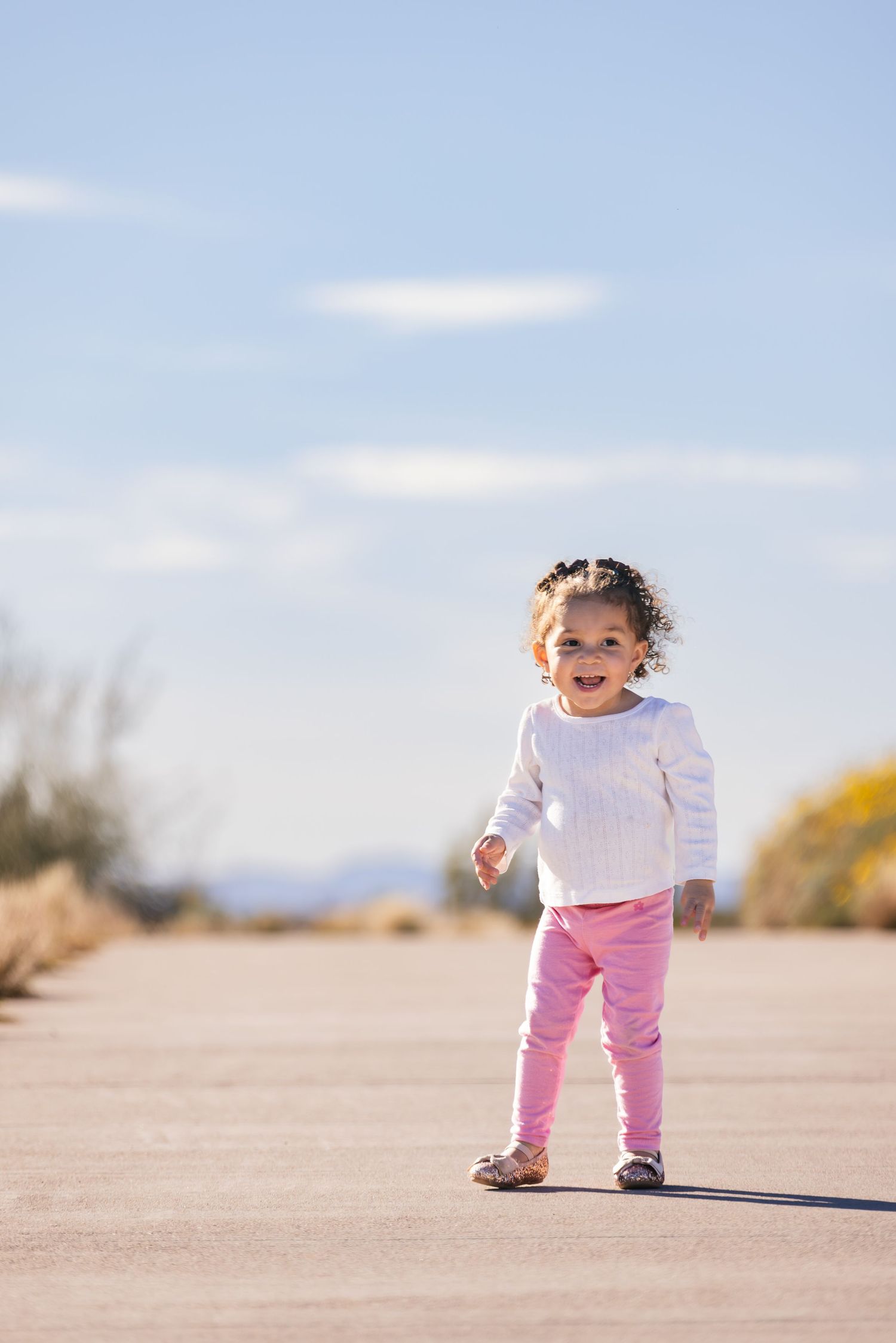 Smiling toddler girl in white sweater and pink leggings standing on a paved path in a sunny desert landscape.