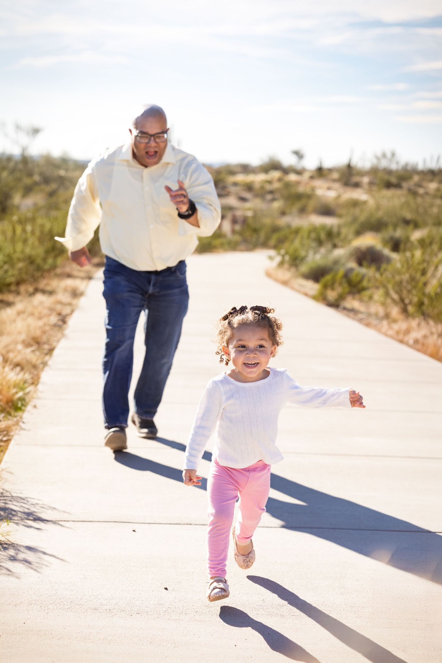 A joyful little girl in pink pants runs ahead of a man on a sunny desert path, both smiling and playing outdoors.