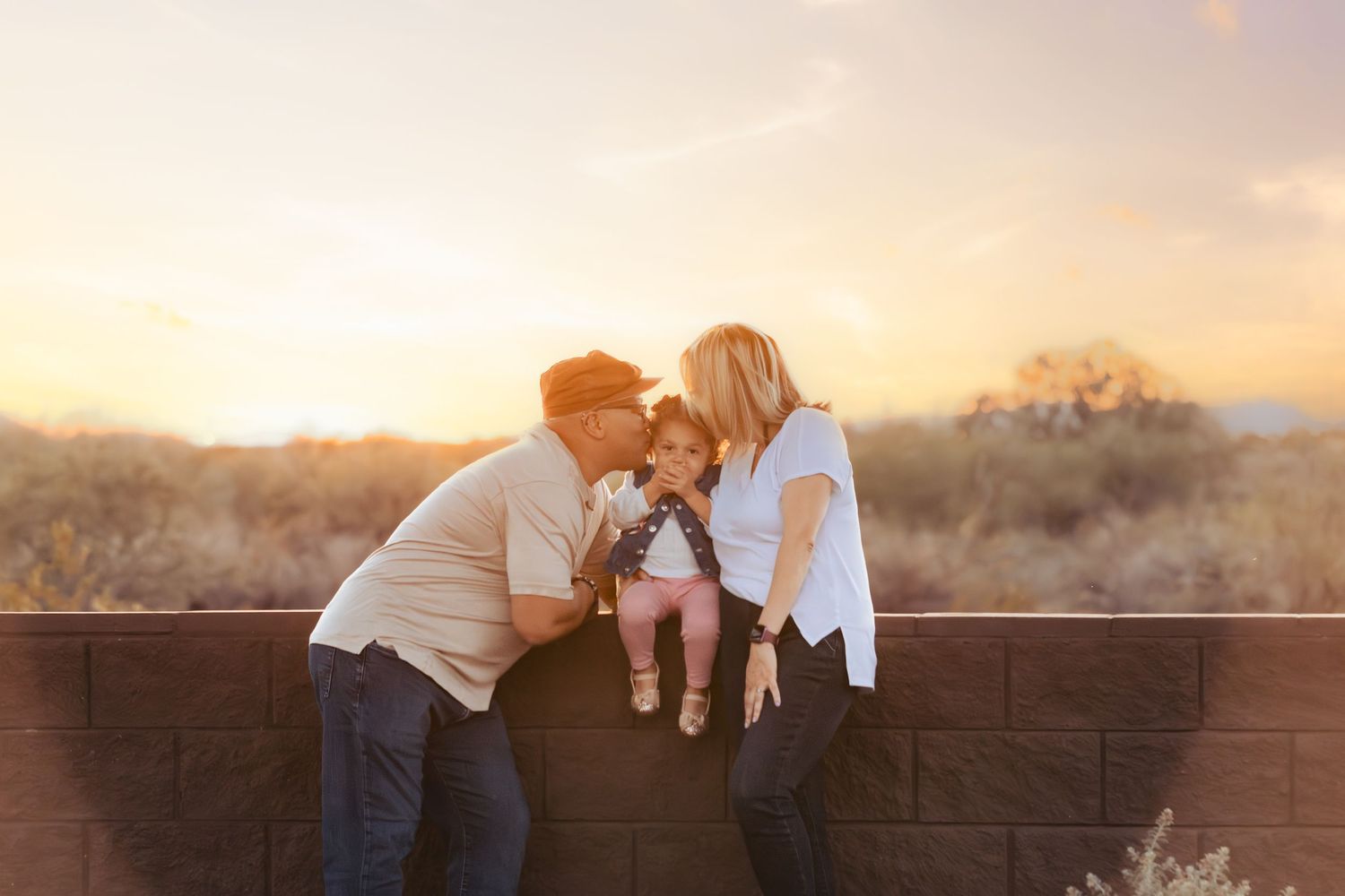 Parents leaning in to kiss their toddler daughter sitting on a brick wall at golden hour sunset.