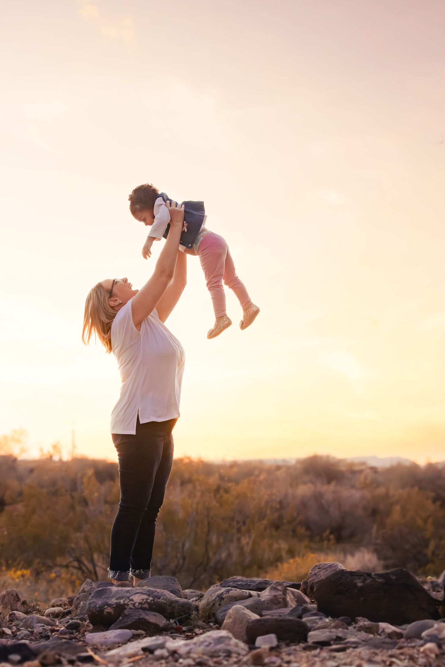 Mother lifting baby joyfully into golden sunset sky outdoors.