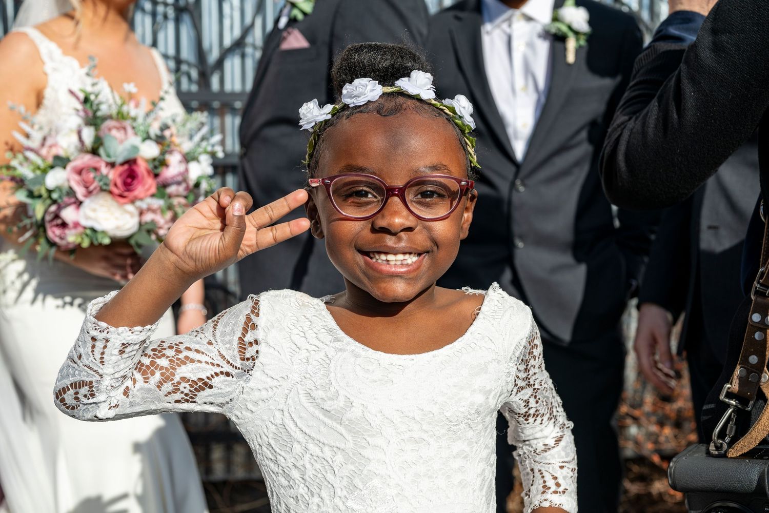 Young flower girl in white lace dress and floral headband smiling and flashing peace sign at wedding ceremony.